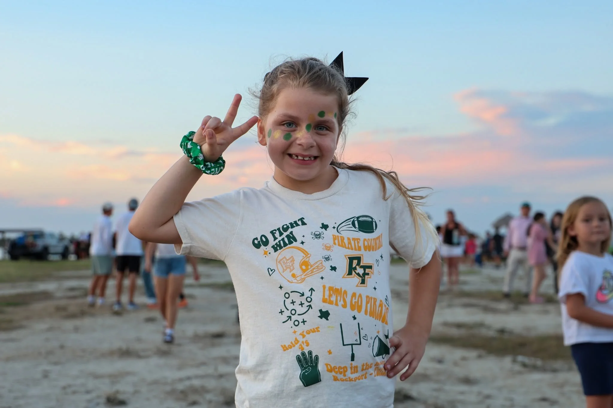 A young girl with face paint and a black bow headband, wearing a white T-shirt with colorful graphics, making a peace sign with her right hand at an outdoor event during sunset.