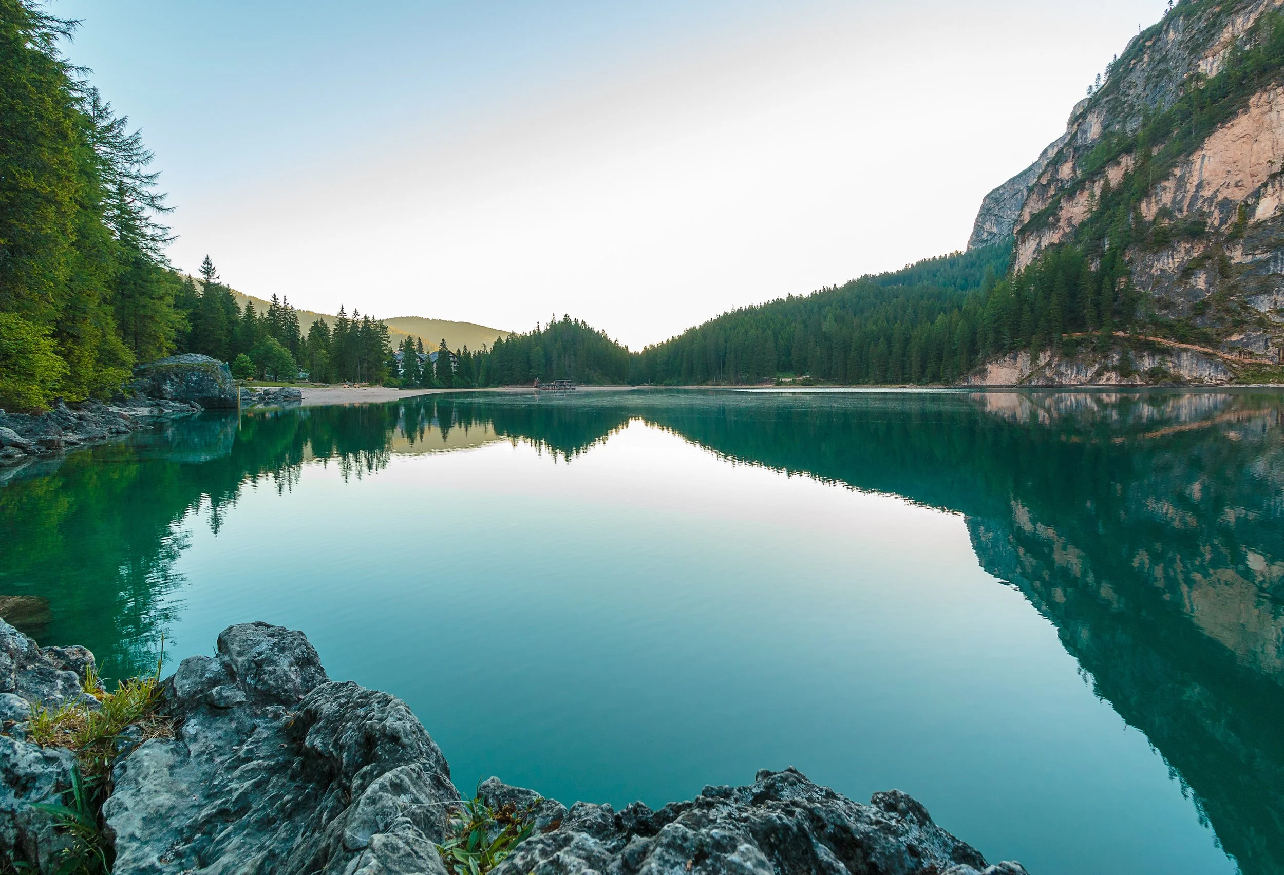 Calm lake surrounded by green pine trees and rocky shoreline, with mountains in the background and a clear sky.