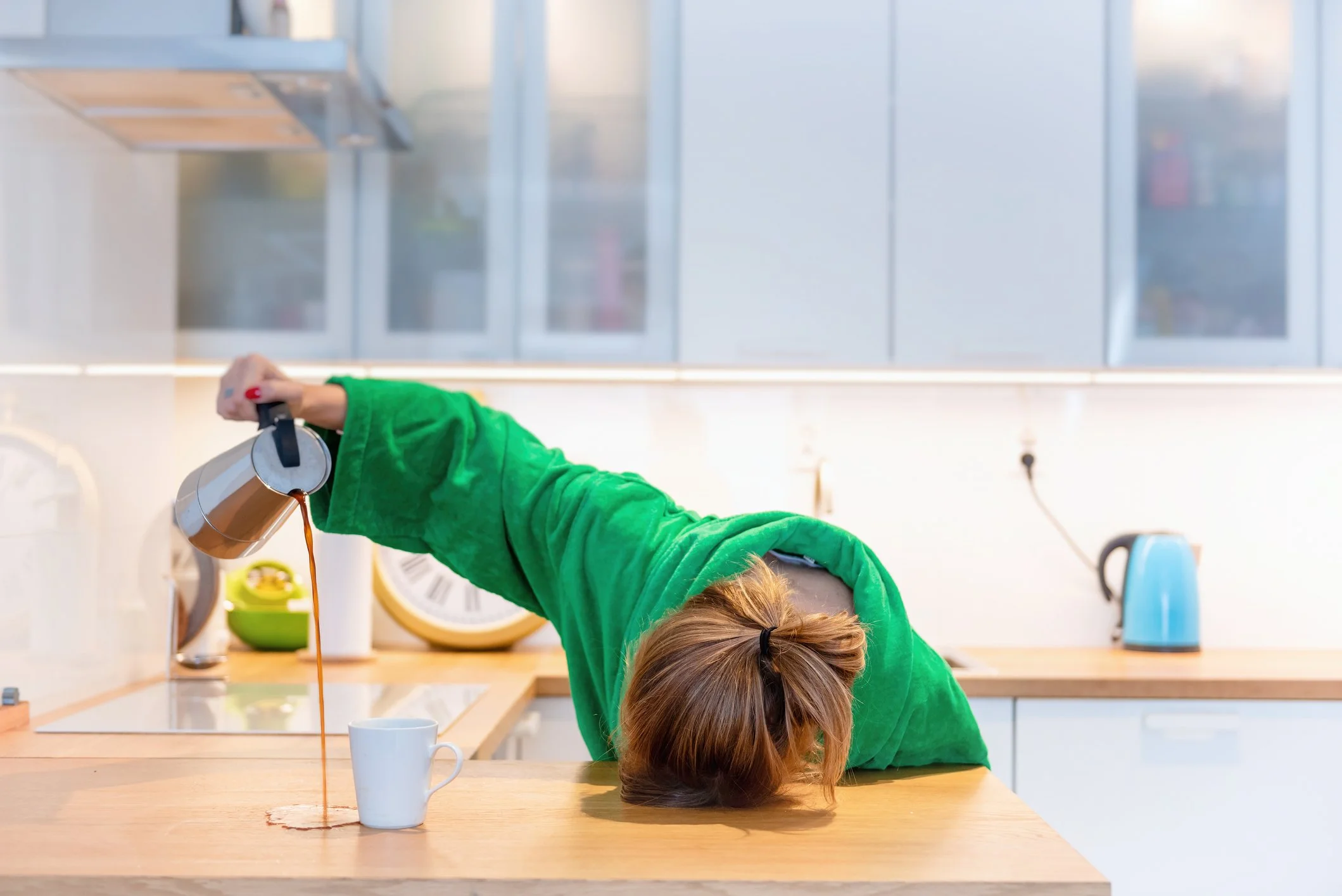 Person in a green hoodie pouring coffee into a mug while another person leans their head on the table in front of them. The kitchen has glass cabinets and a wooden countertop.