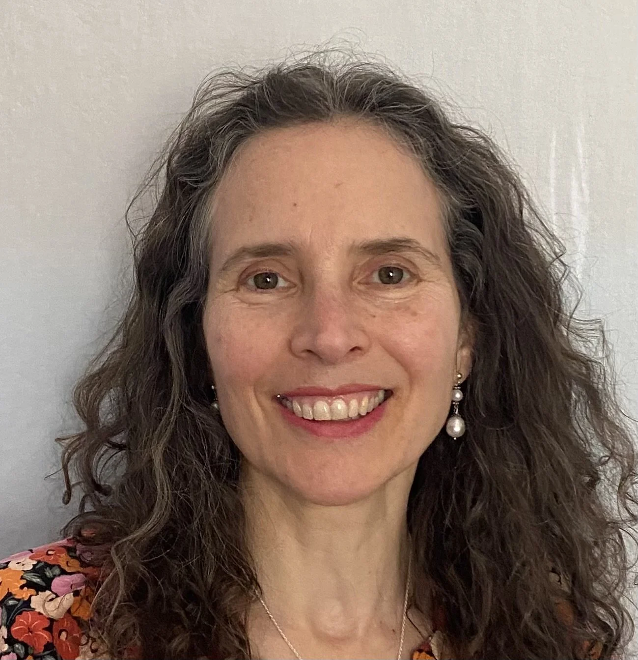 A woman with curly brown hair, smiling, wearing pearl earrings and a floral top.
