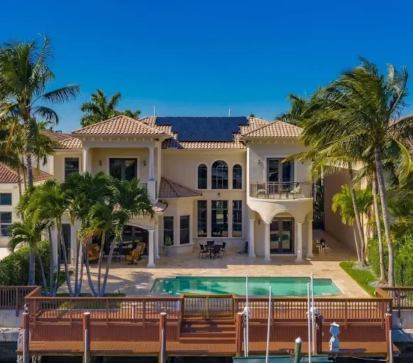 Backyard with swimming pool and luxury house surrounded by palm trees under a blue sky.