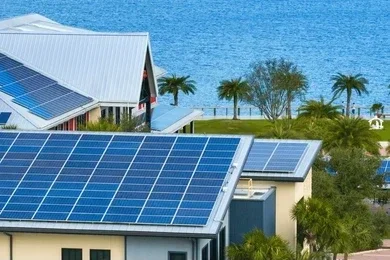 Residential buildings with solar panels on rooftops near a body of water, surrounded by palm trees.