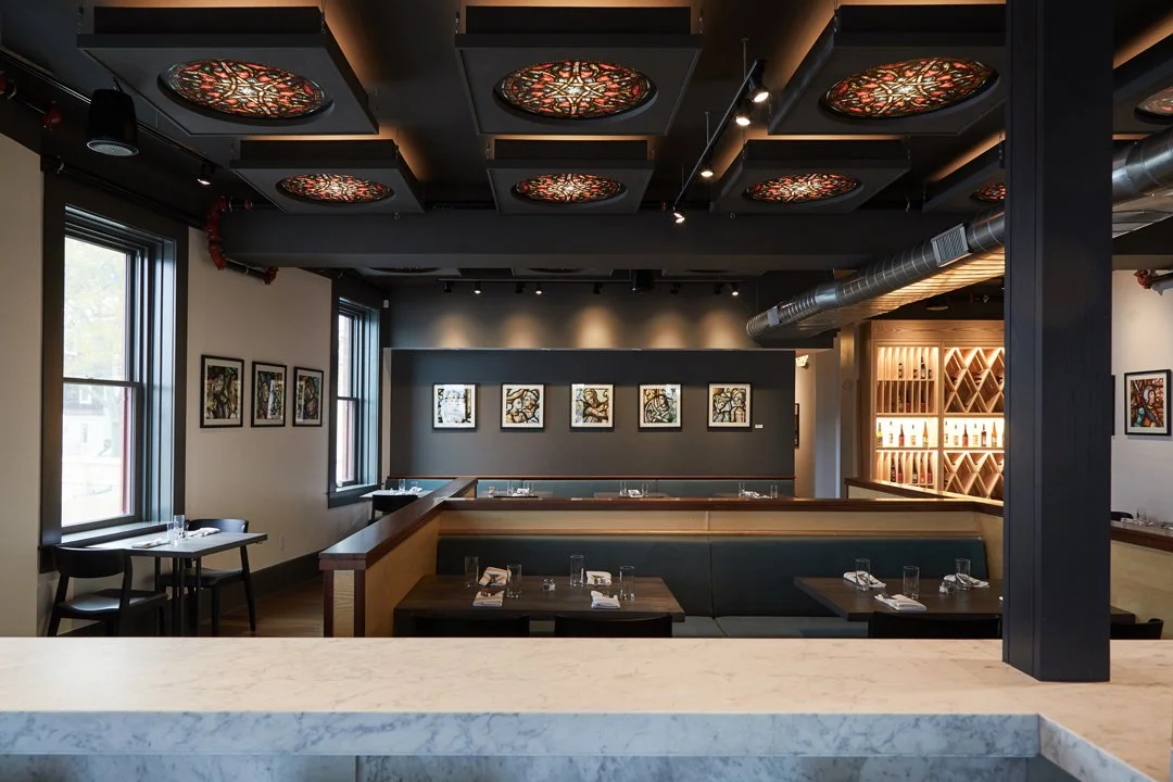 Interior of a modern restaurant with art on the walls, large windows, and decorative ceiling panels.