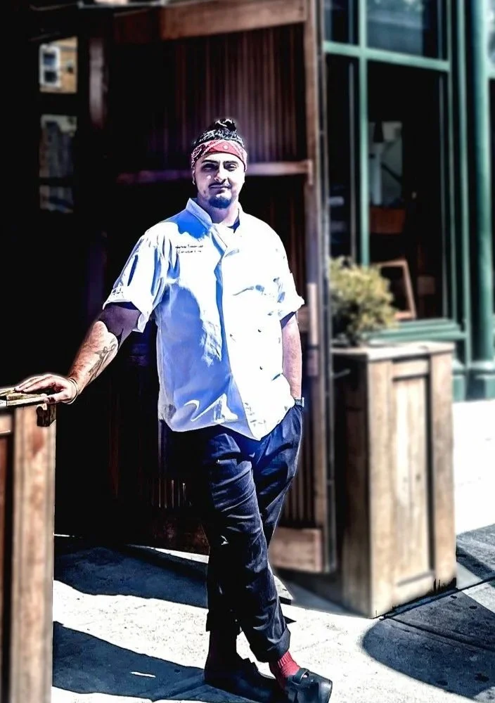 A man wearing a chef's uniform leaning against a wooden counter outside, with wooden and green structures in the background.