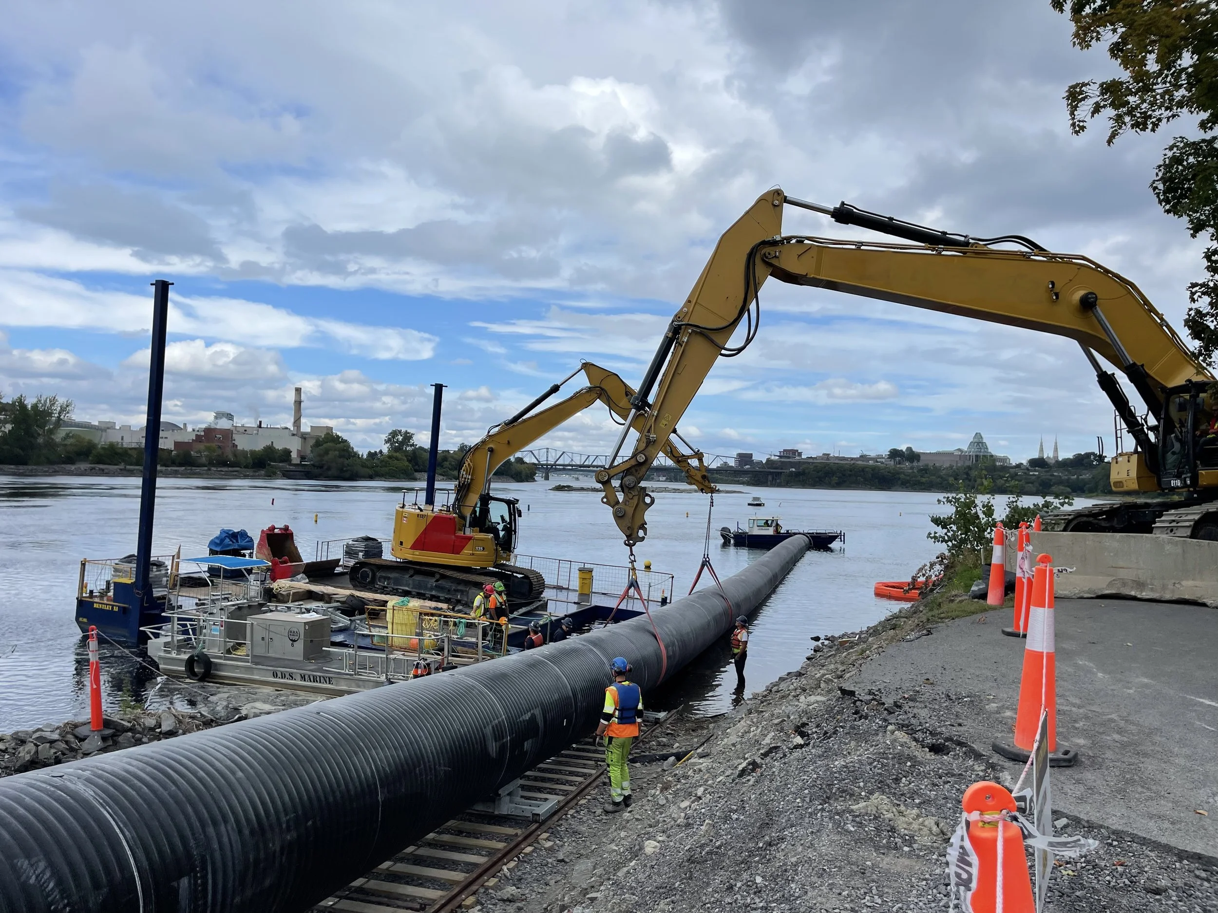 Workers using excavators to install a large pipeline along a riverside during daytime, with a cloudy sky.