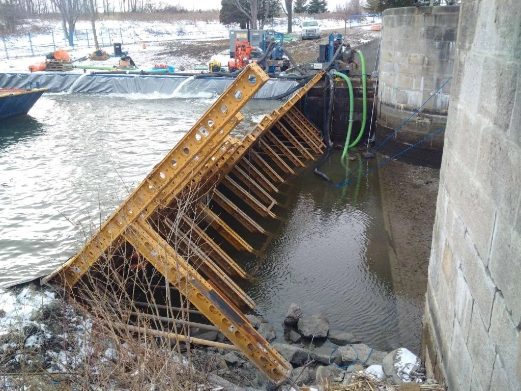 A collapsed cofferdam with yellow metal sheets at a waterway, with construction and machinery in the background and snow on the ground.