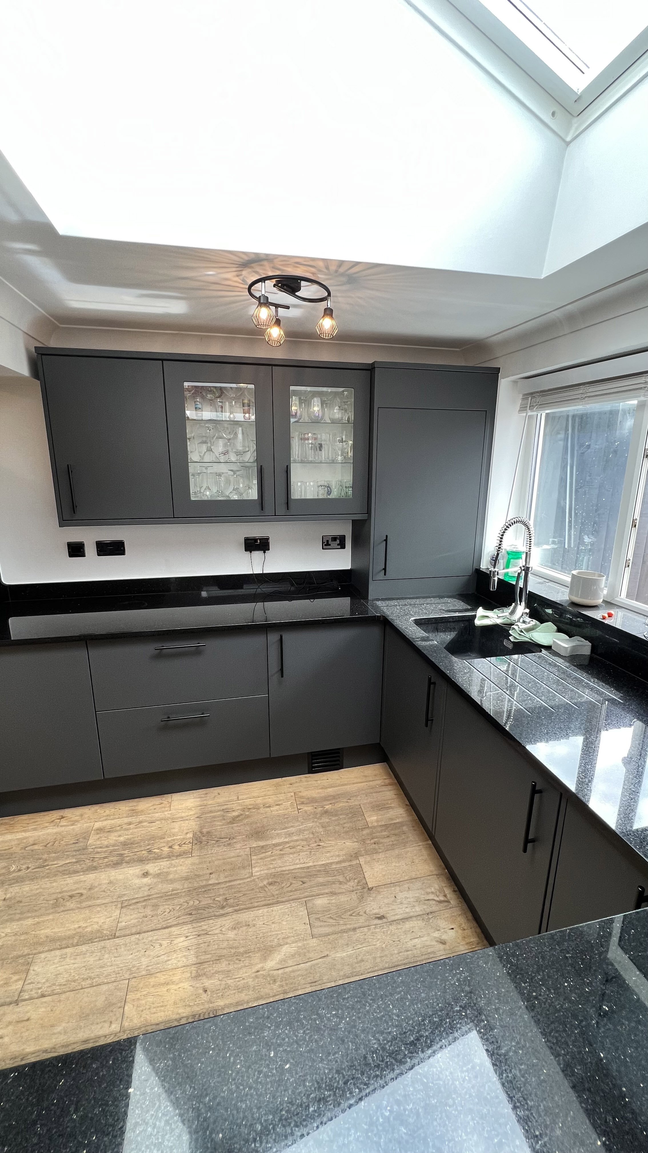 Modern kitchen interior with dark gray cabinets, black granite countertops, wooden flooring, and a skylight. Features glass cabinet doors, a sleek faucet, and under-cabinet lighting.