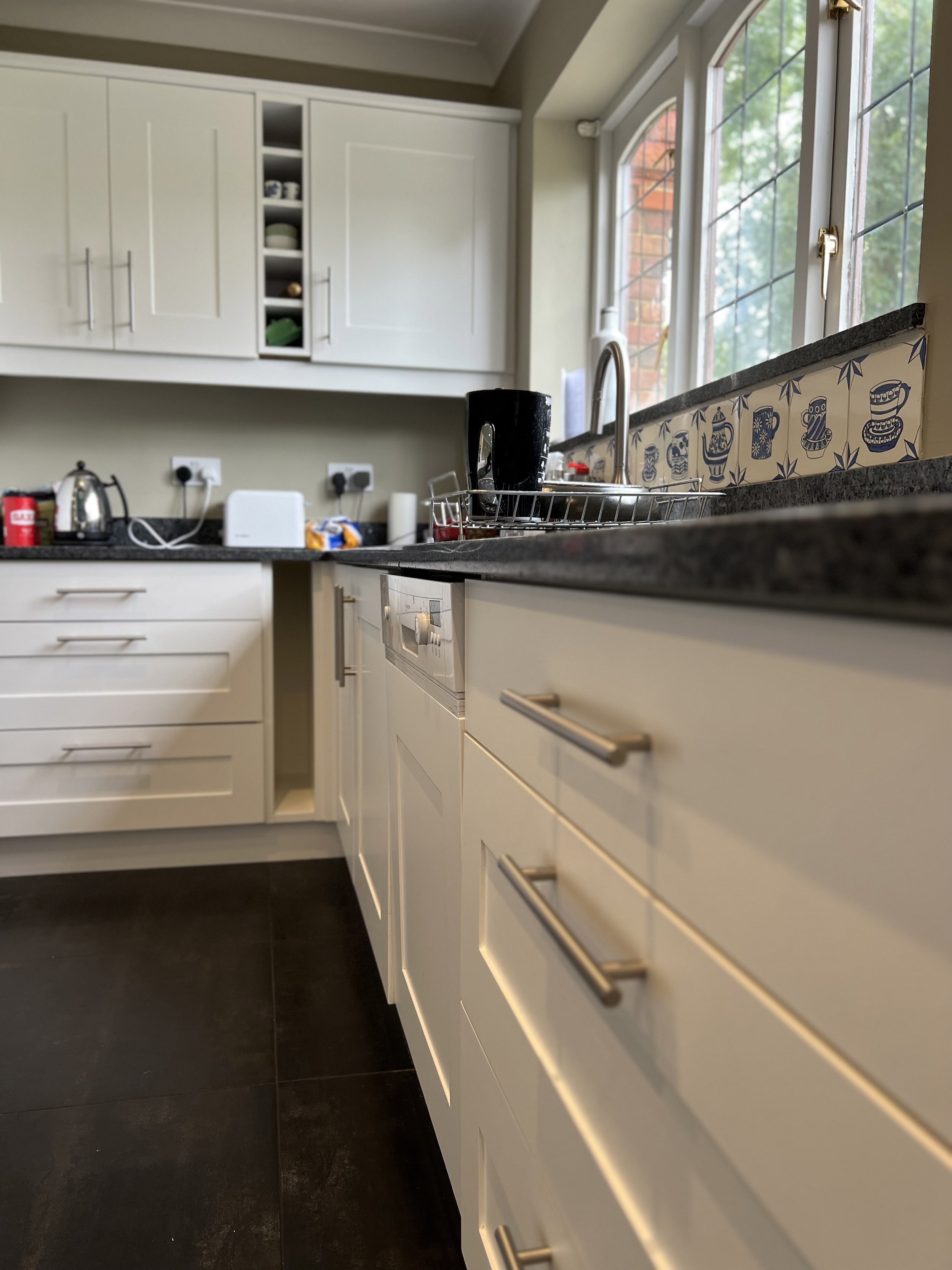 Modern kitchen with white cabinetry, black countertop, stainless steel handles, and a tiled backsplash featuring blue designs. A window with a garden view is visible above the sink.