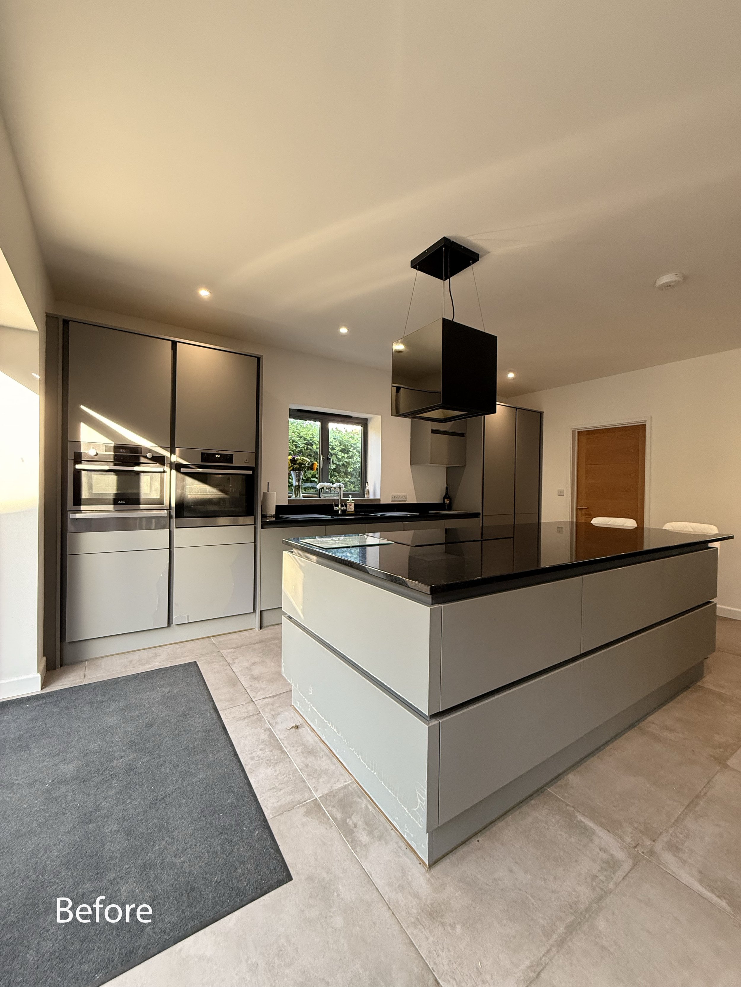 Modern kitchen with gray cabinetry, large central island, built-in appliances, tiled floor, and overhead black range hood. "Before" caption on image.