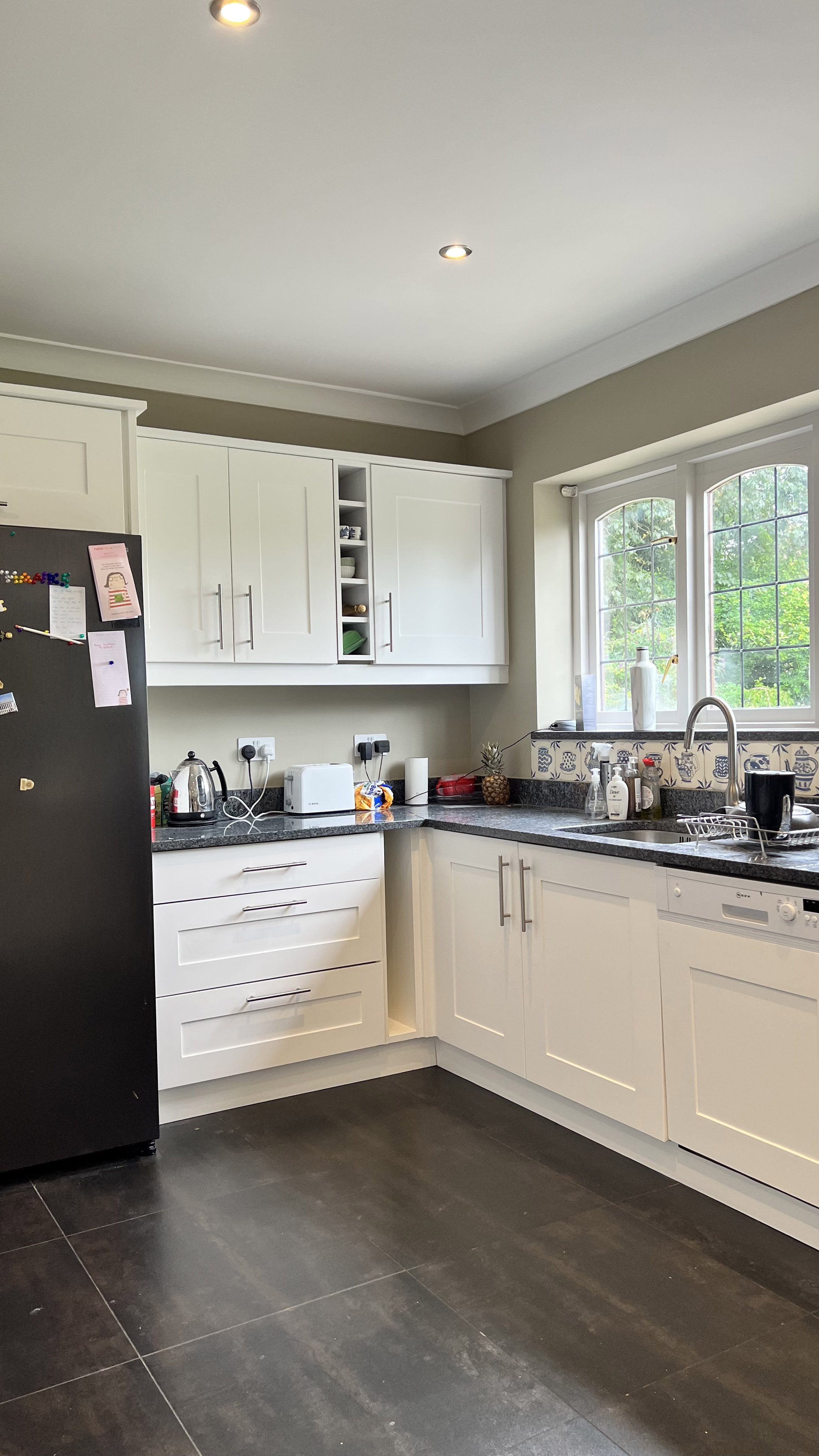 Modern kitchen with black refrigerator, white cabinets, gray countertops, and a window above the sink with a view of greenery. Appliances include a stainless steel kettle, toaster, and a white dishwasher. A pineapple and bread are also on the counter