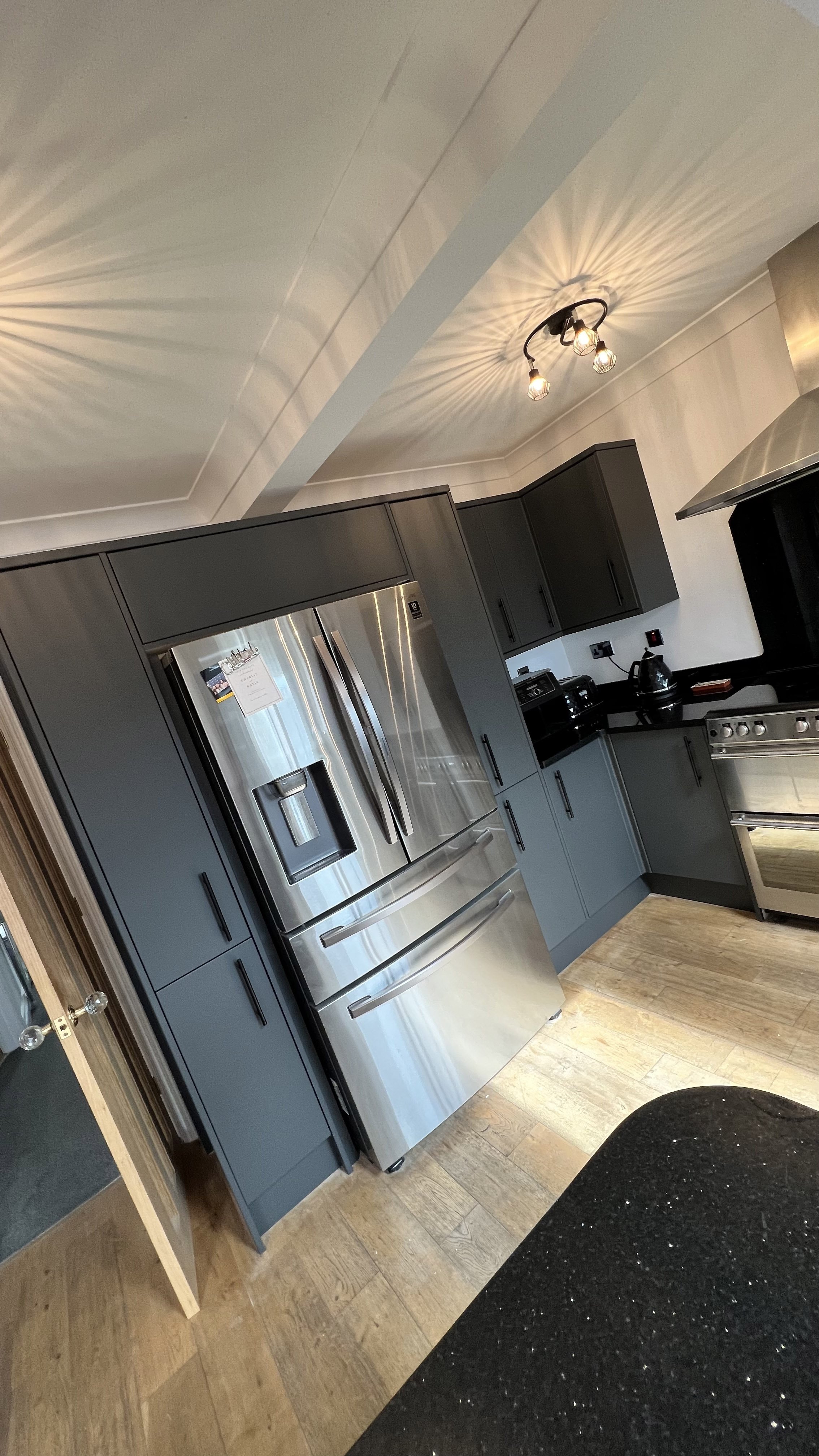 Modern kitchen with stainless steel appliances, including a French door refrigerator, dark cabinetry, wooden floor, and contemporary lighting fixture.