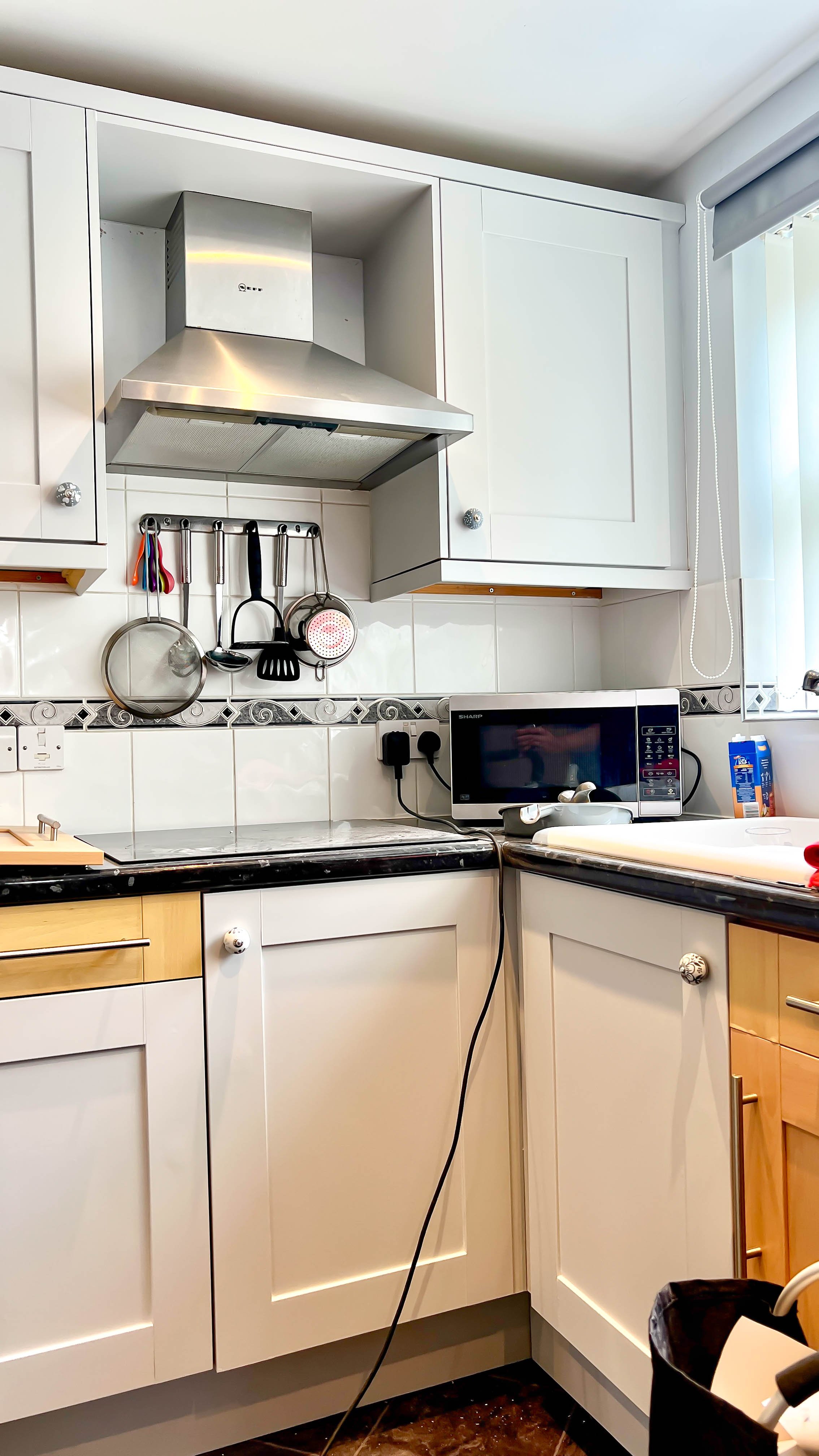 A modern kitchen with white cabinets, a stainless steel range hood, hanging utensils, a microwave, and a countertop with a cutting board. The walls are tiled and there is a window with blinds.