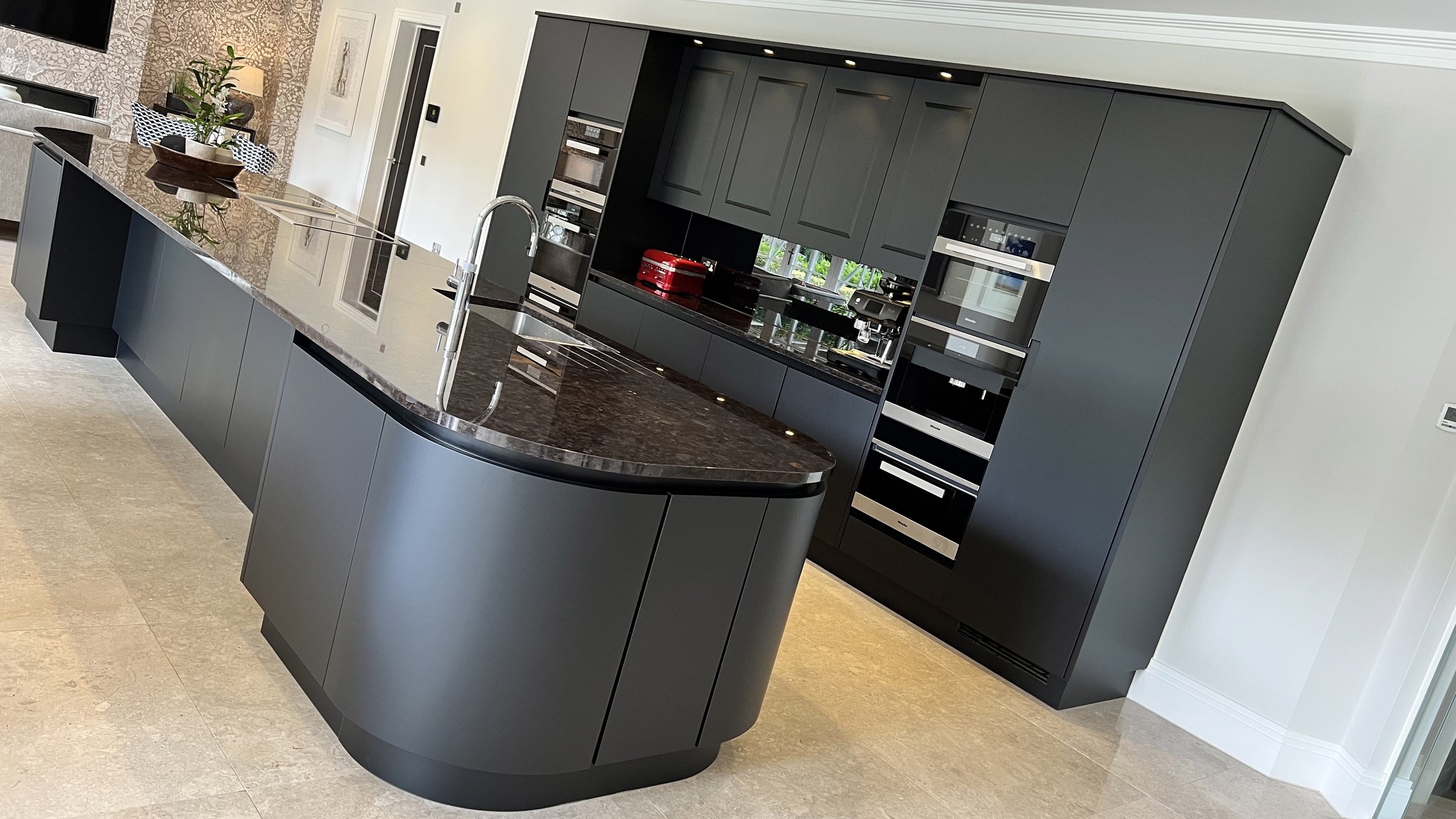 Modern kitchen with sleek black cabinetry, stainless steel appliances, and a large curved island with a dark countertop and a built-in sink.