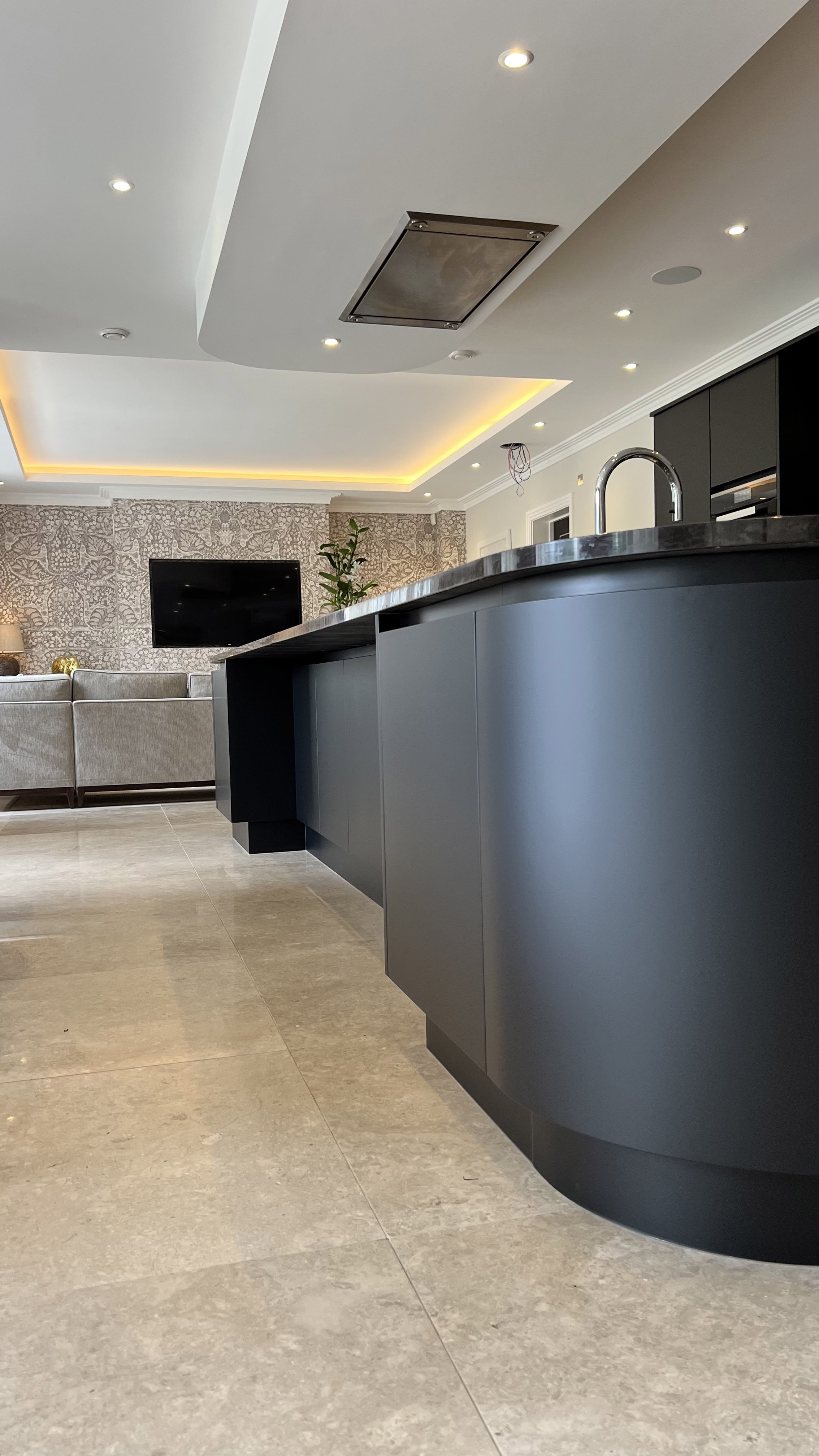 Modern kitchen with a large dark-colored island, marble countertops, stainless steel faucet, recessed lighting, and a patterned wallpaper in the background.