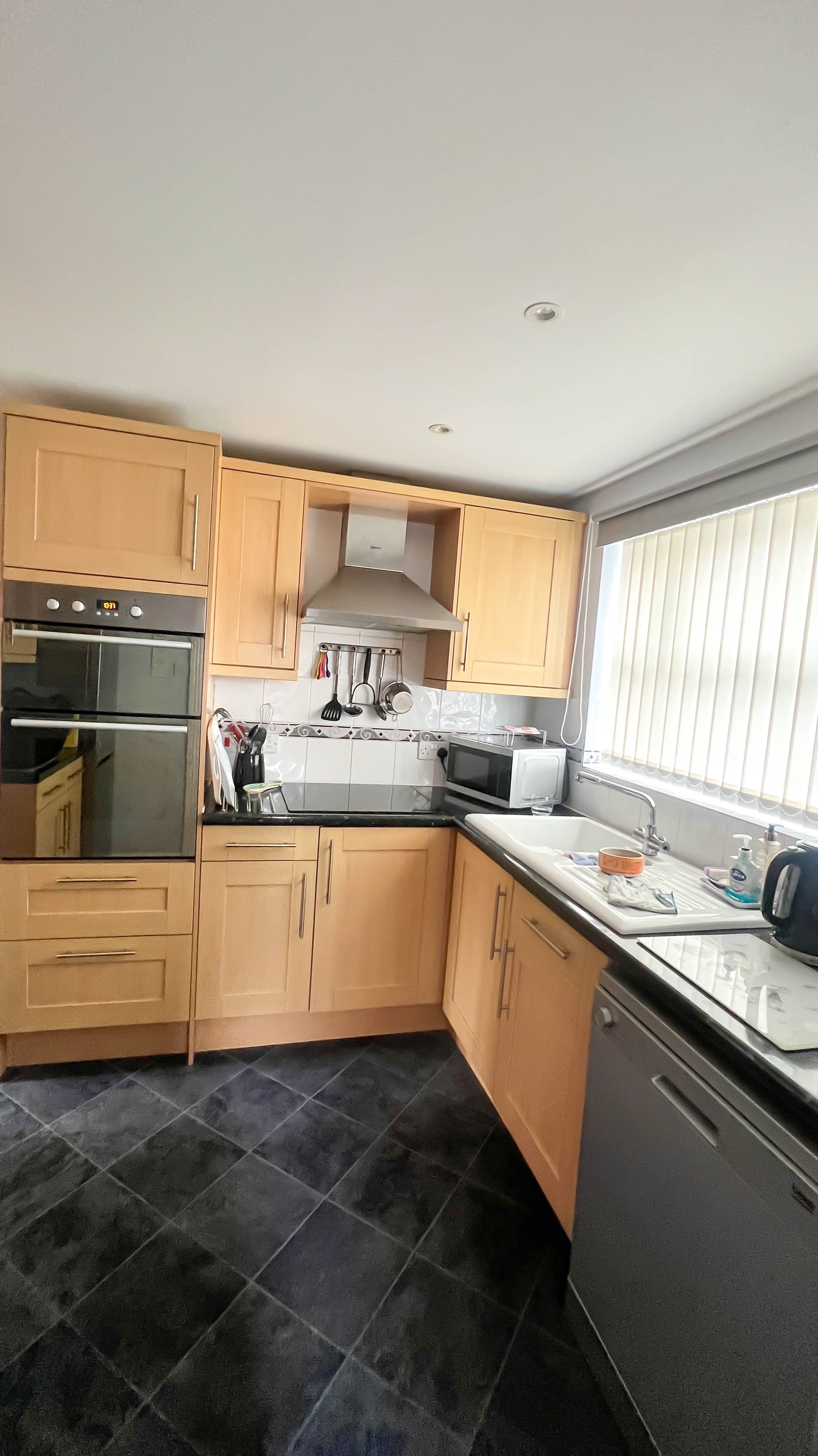 Modern kitchen with wooden cabinets, double oven, stove, microwave, sink, and utensils, featuring a tiled floor and window blinds.