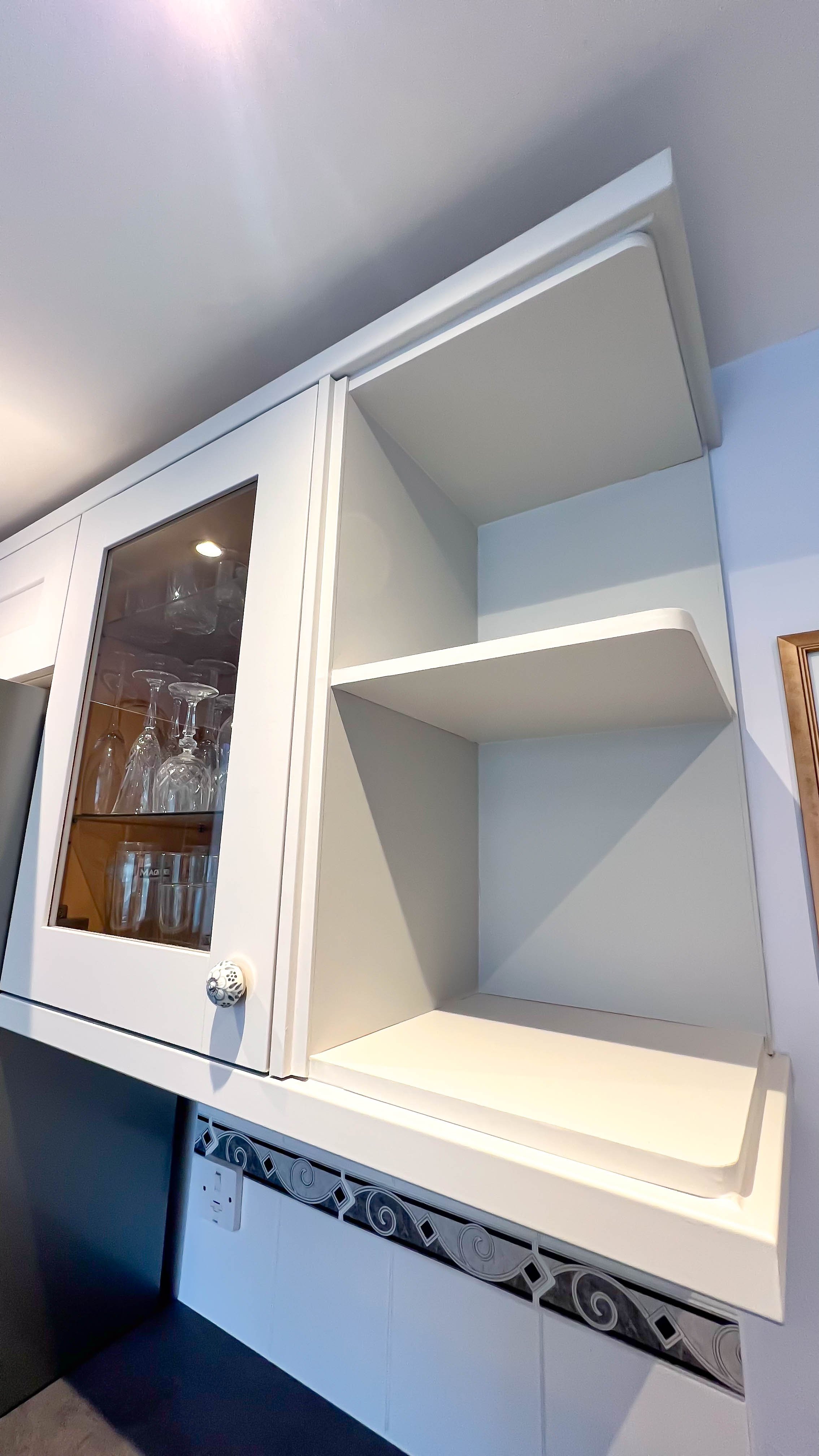 White kitchen cabinet with glass door, displaying wine glasses and other glassware inside. Adjacent open corner shelves are empty. Decorative tile below.