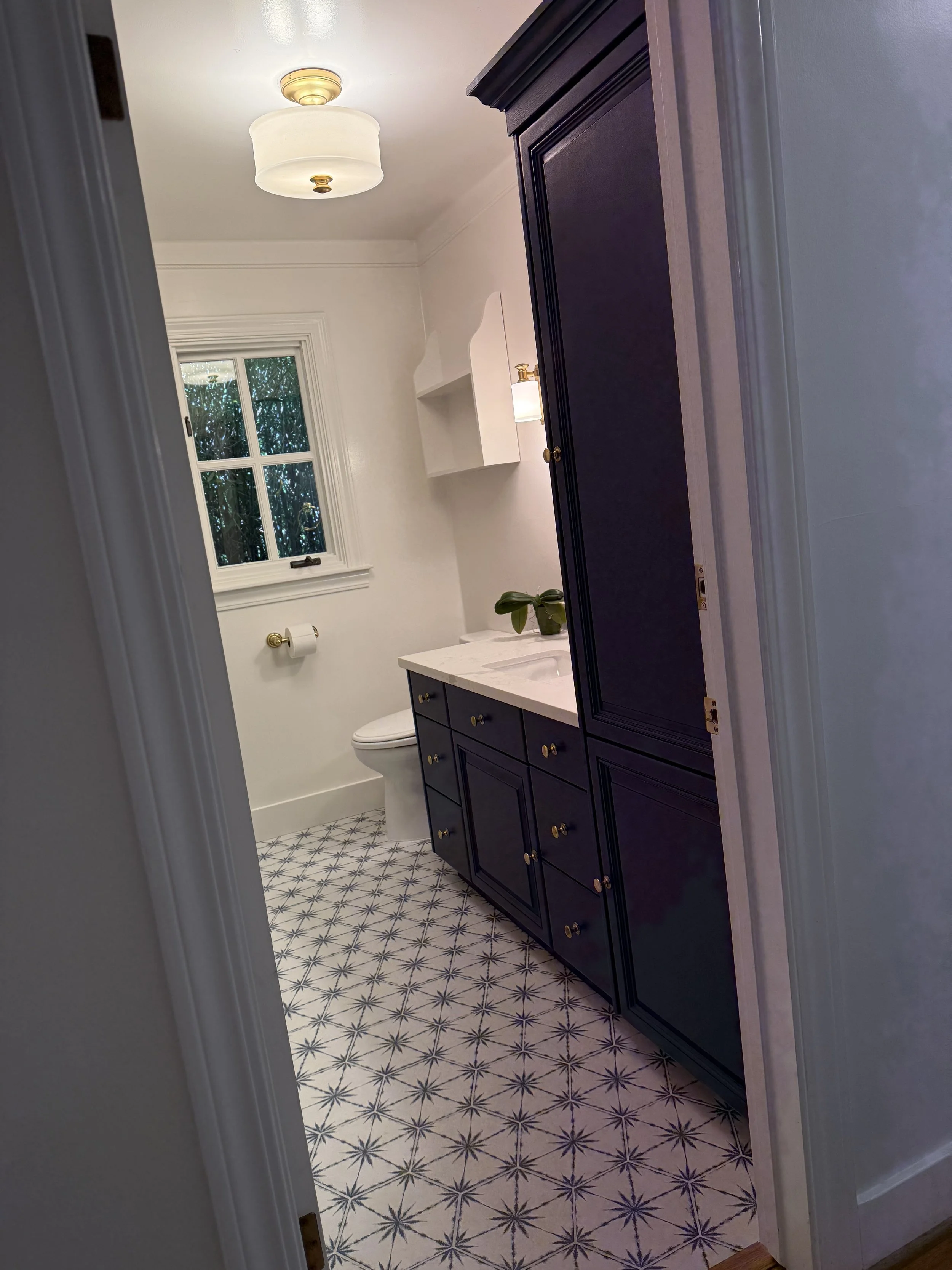 View of a small bathroom with a black cabinet, white marble countertop, a white toilet, a small window showing greenery outside, and patterned tile flooring. There is a ceiling light fixture and a wall-mounted light above the sink.