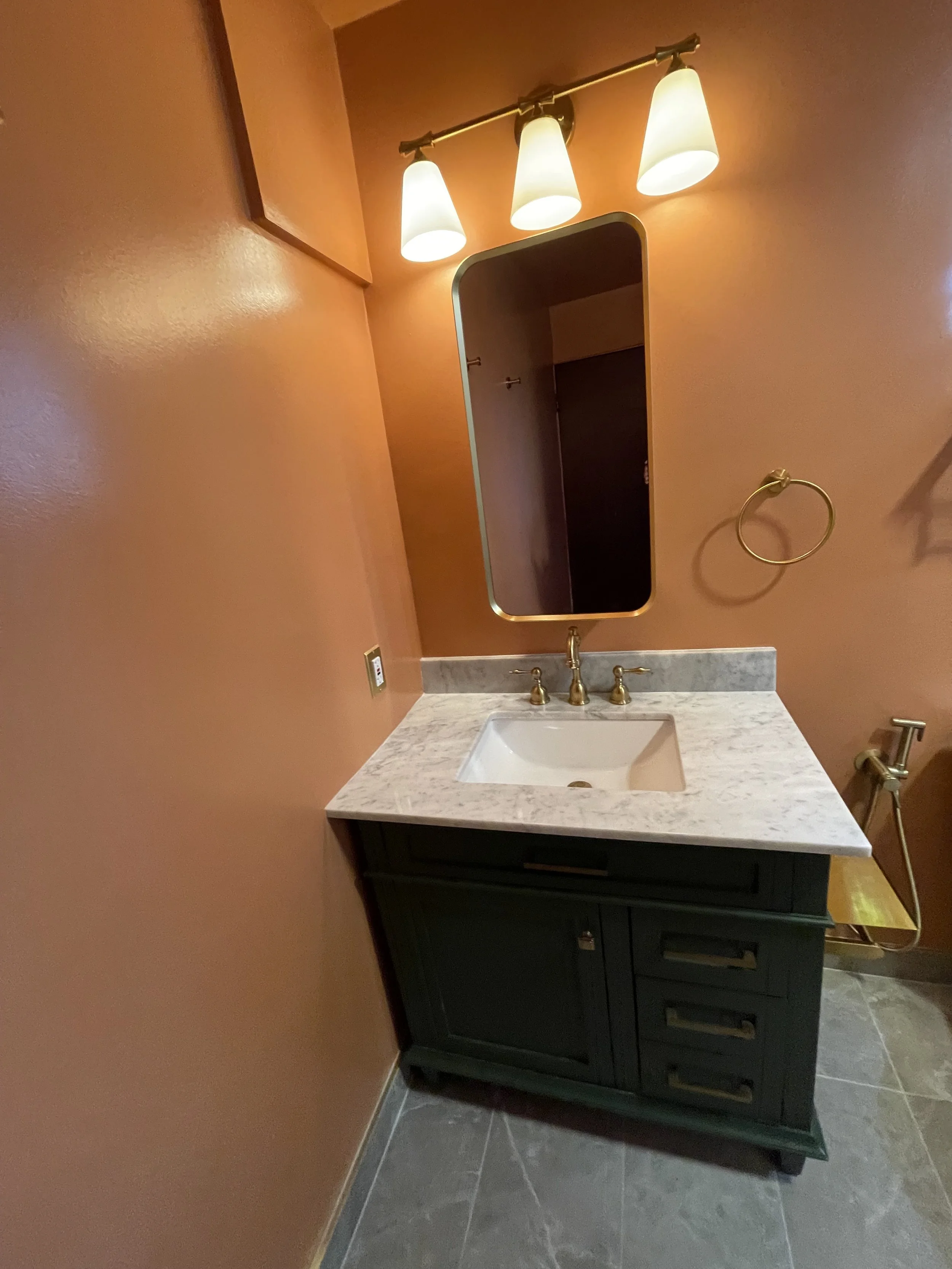 Bathroom with orange painted walls, a rectangular mirror with a gold frame, three light fixtures above, a white marble countertop with a white sink, and brass fixtures. There are also towel rings and a yellow towel on the side.