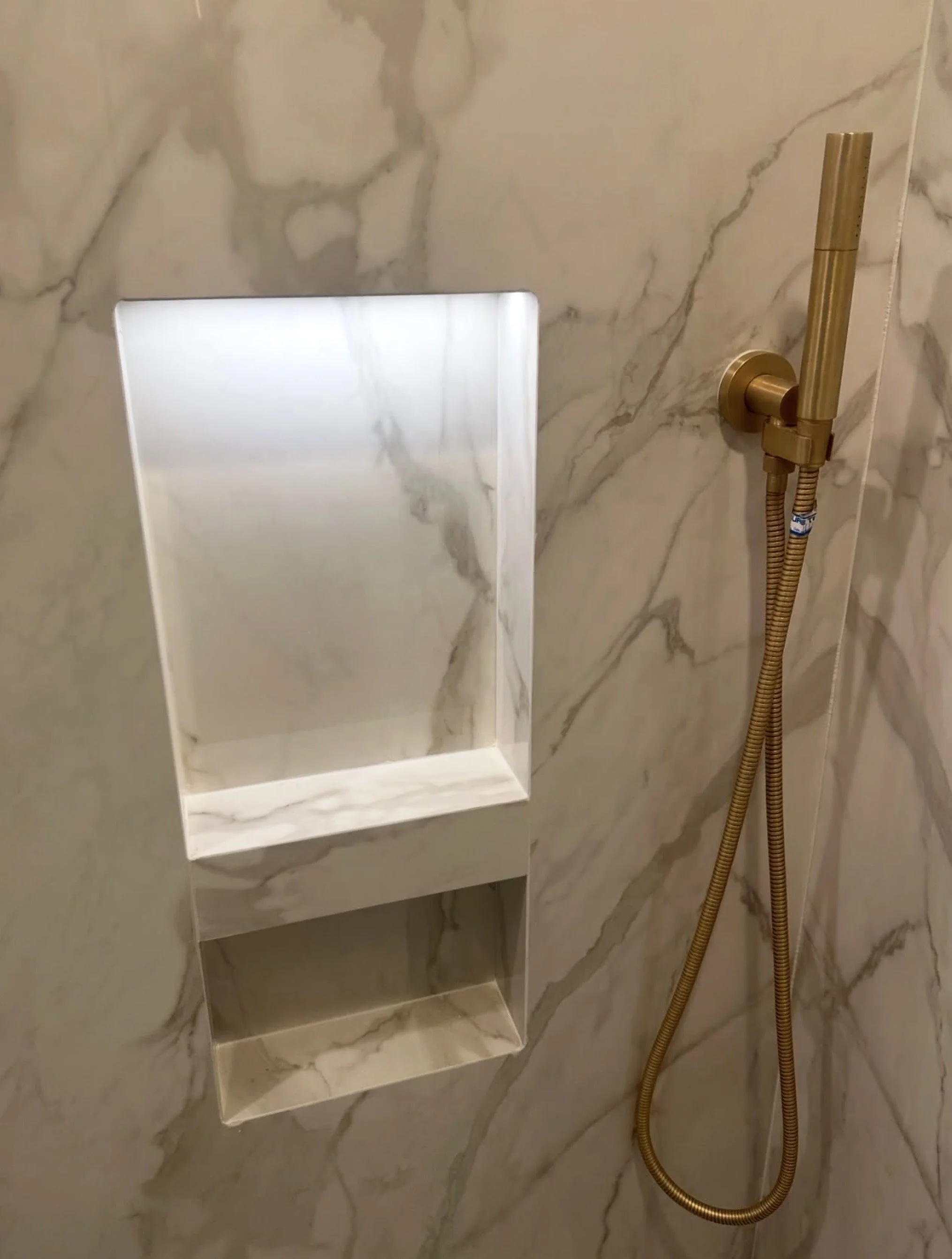 Marble shower wall with built-in mirror and gold handheld showerhead.