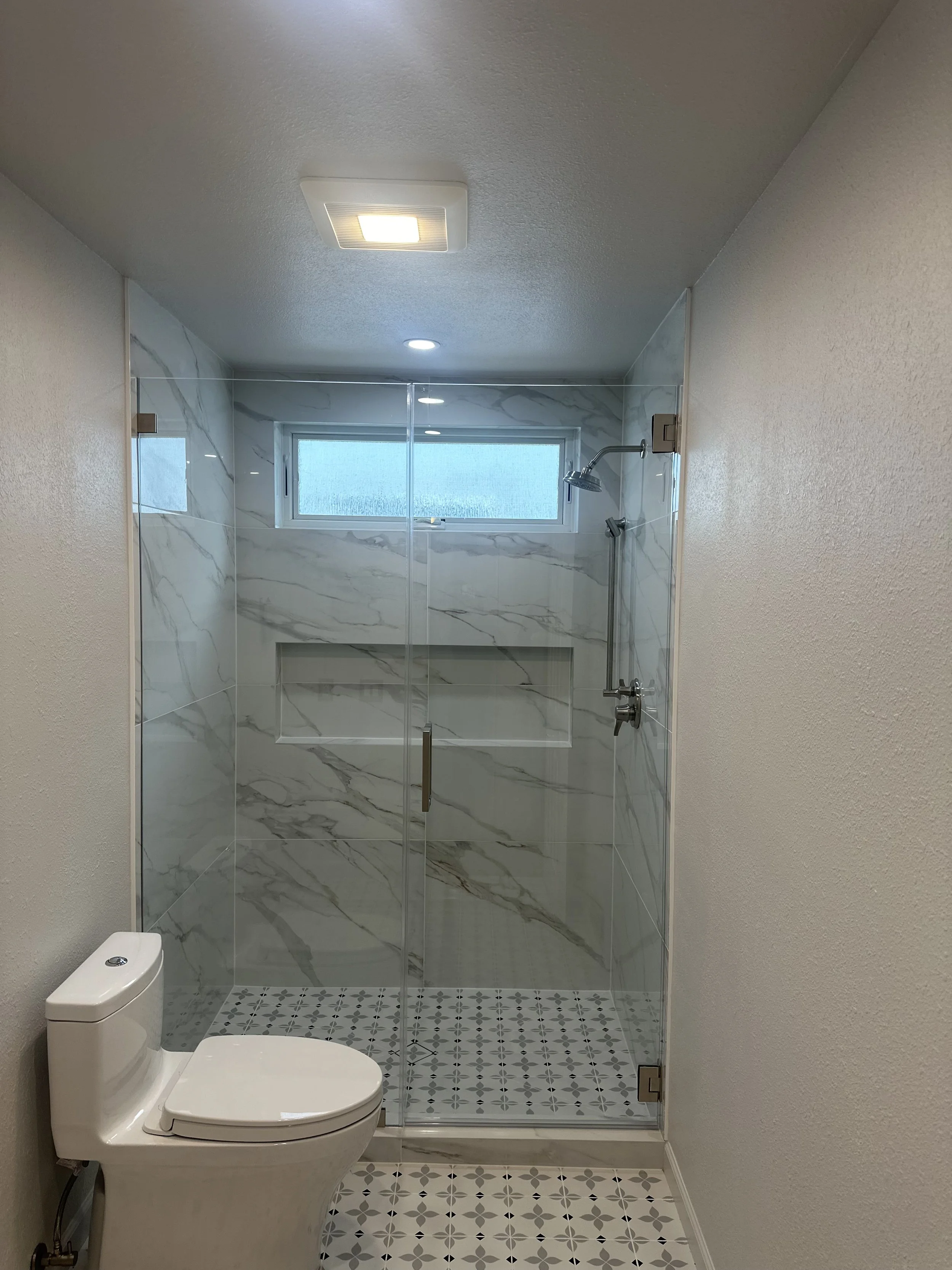 A modern bathroom with a walk-in shower enclosed by clear glass doors, white marble-patterned tiles, a small rectangular window, a ceiling-mounted light, and a white toilet in front of the shower.