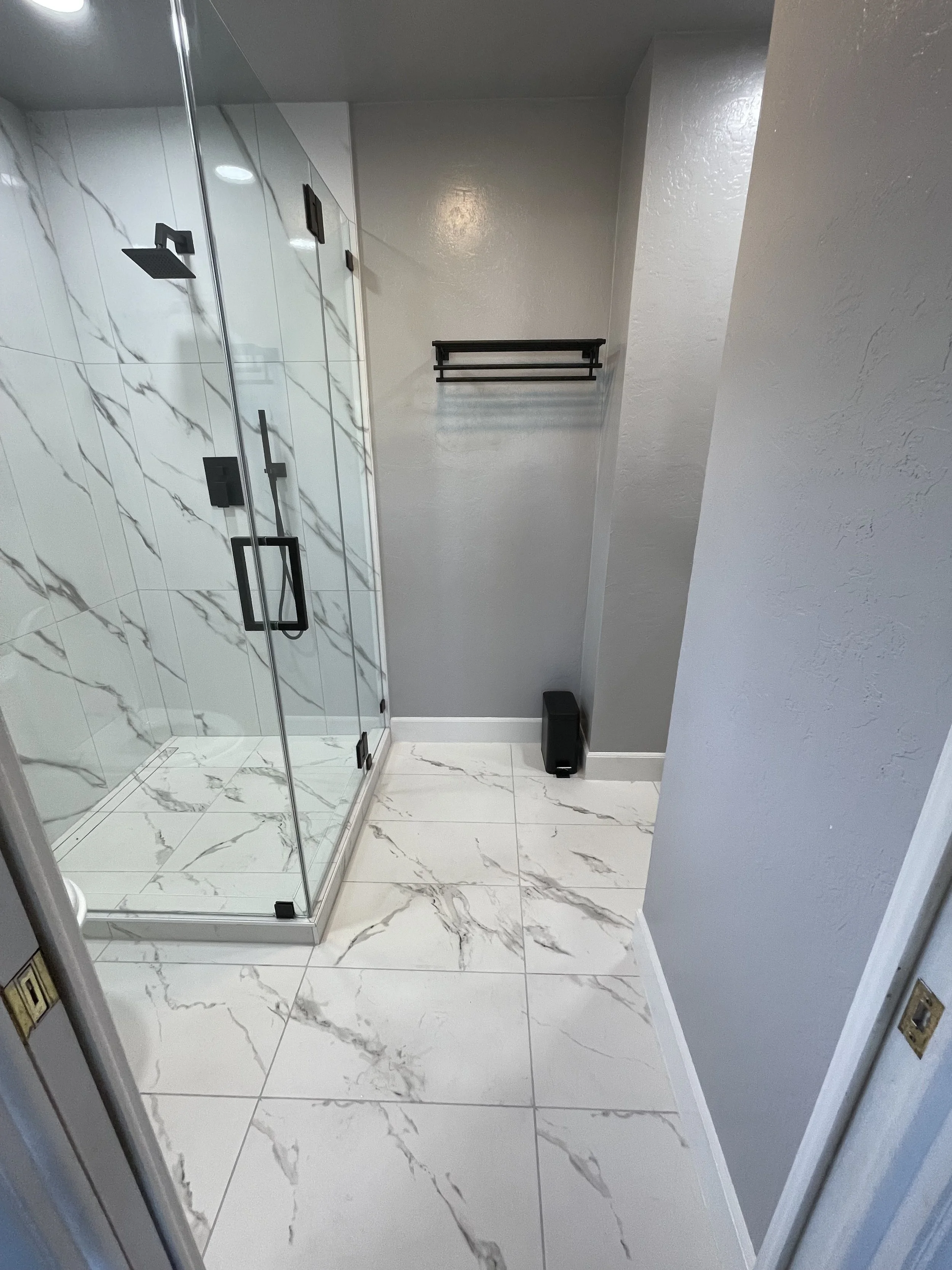 A modern bathroom with a glass shower enclosure featuring black fixtures, gray walls, and white marble floor tiles with gray veining.
