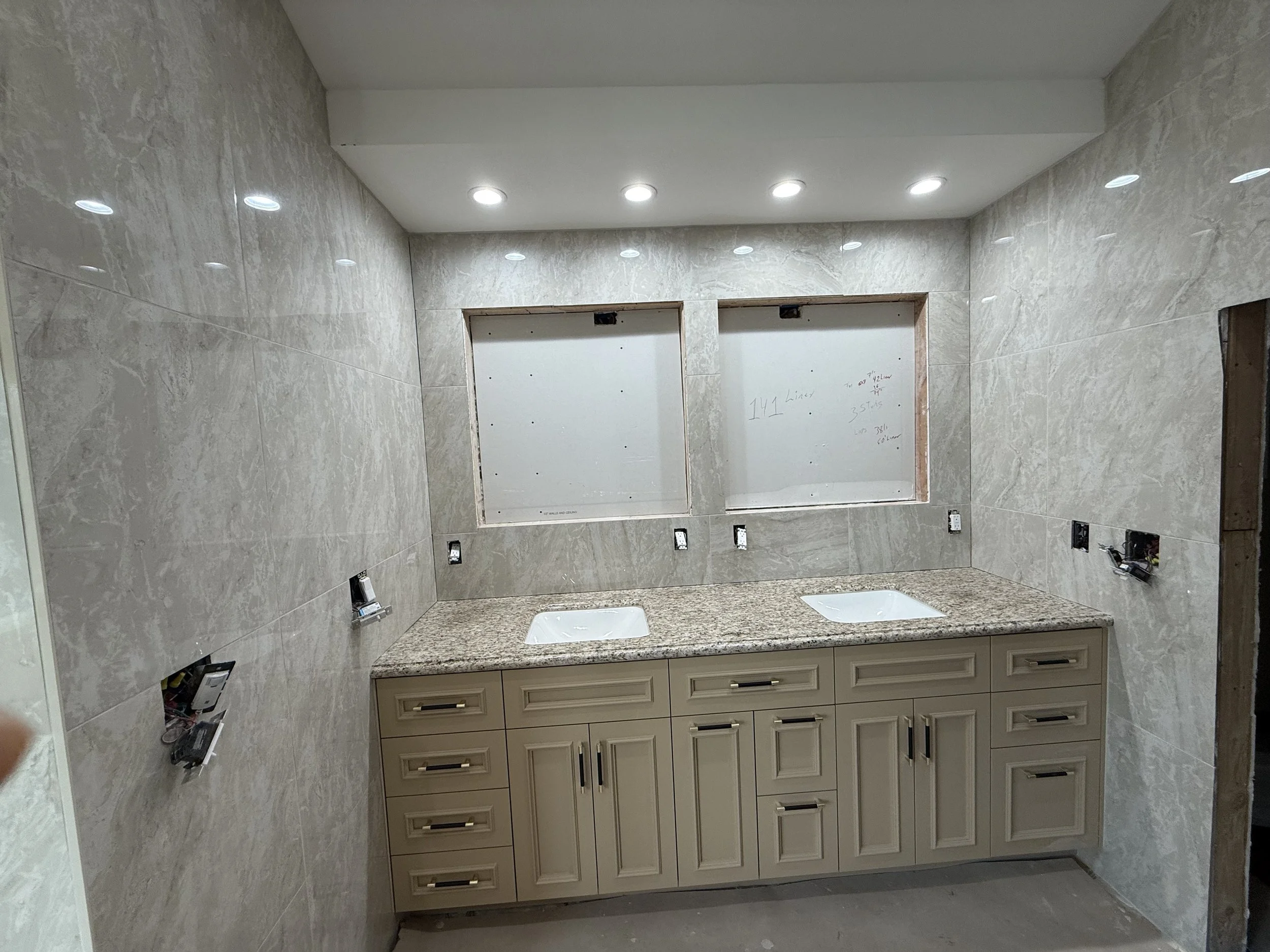 Bathroom under construction with tiled walls, a granite countertop with two built-in sinks, cream-colored cabinets, and no faucets installed. Two unfinished window openings are covered with white boards, and there are electrical outlets and wiring vi