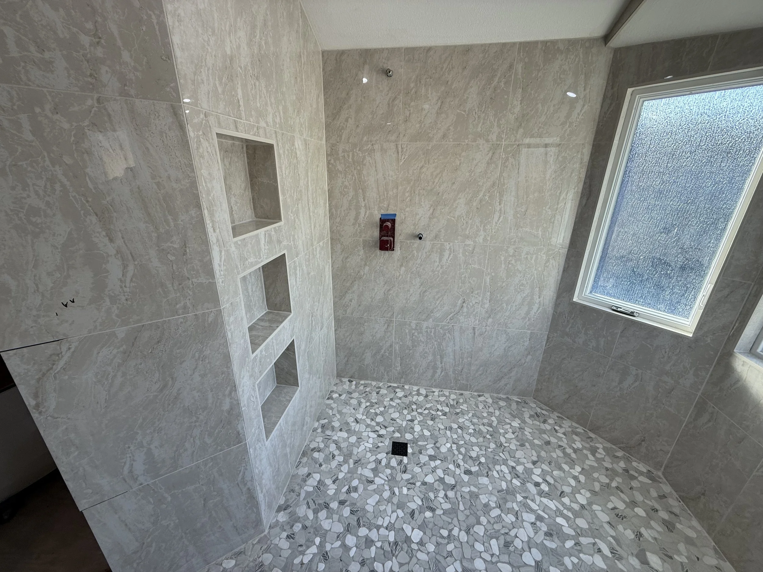 A tiled bathroom shower with beige marble walls, a mosaic pebble floor, built-in wall niches, a window with frosted glass, and an overhead showerhead mount.