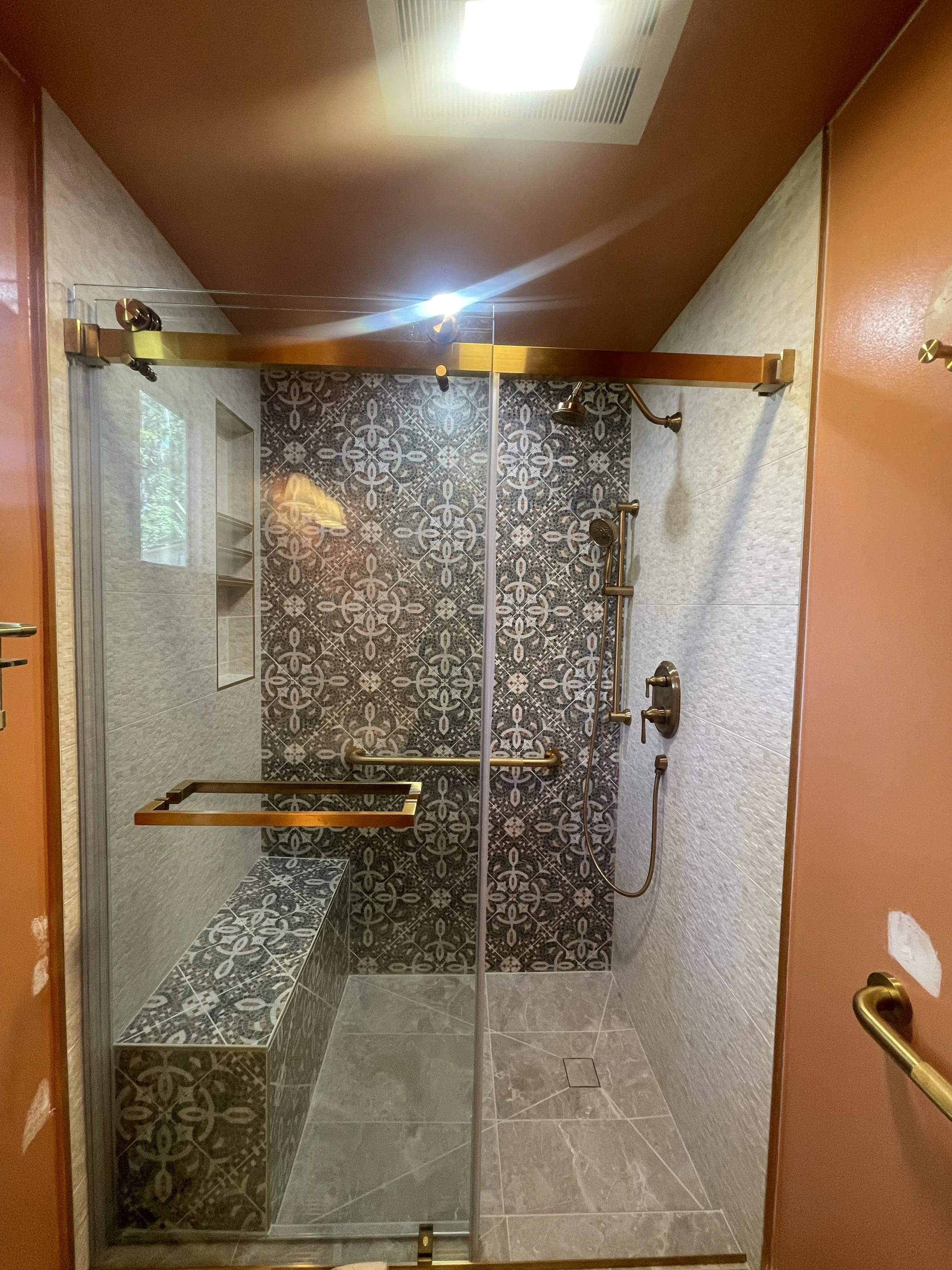 A walk-in shower with glass door, decorative patterned tiles, a built-in bench, a showerhead, and a handheld shower on a sliding bar, with bronze fixtures and a ceiling light.