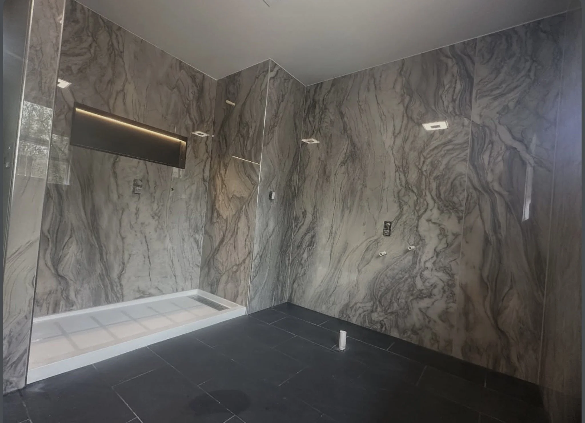 Empty modern bathroom with marble walls, black floor tiles, and construction fixtures, including a vent on the floor and wall, electrical outlets, white pipe on the floor, and partial mirrors.
