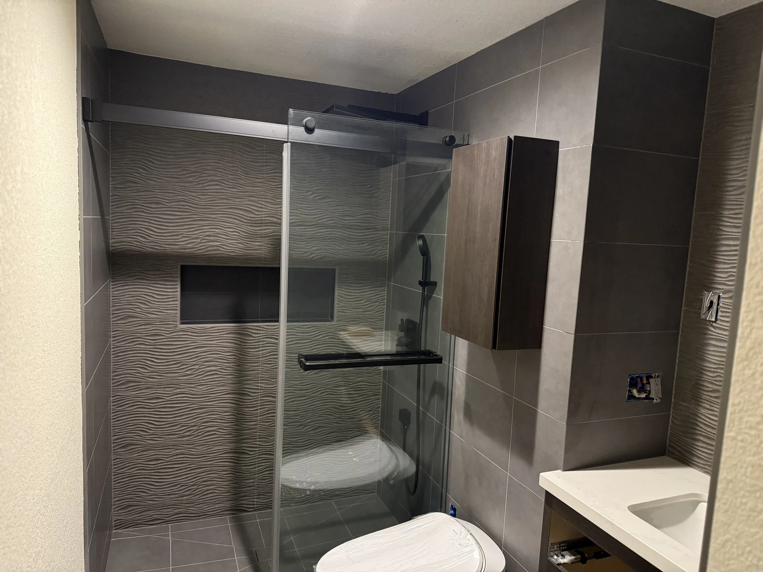 Modern bathroom with a glass-enclosed shower, textured wall tiles, a wall-mounted dark wood cabinet, a small sink, and an electrical outlet.