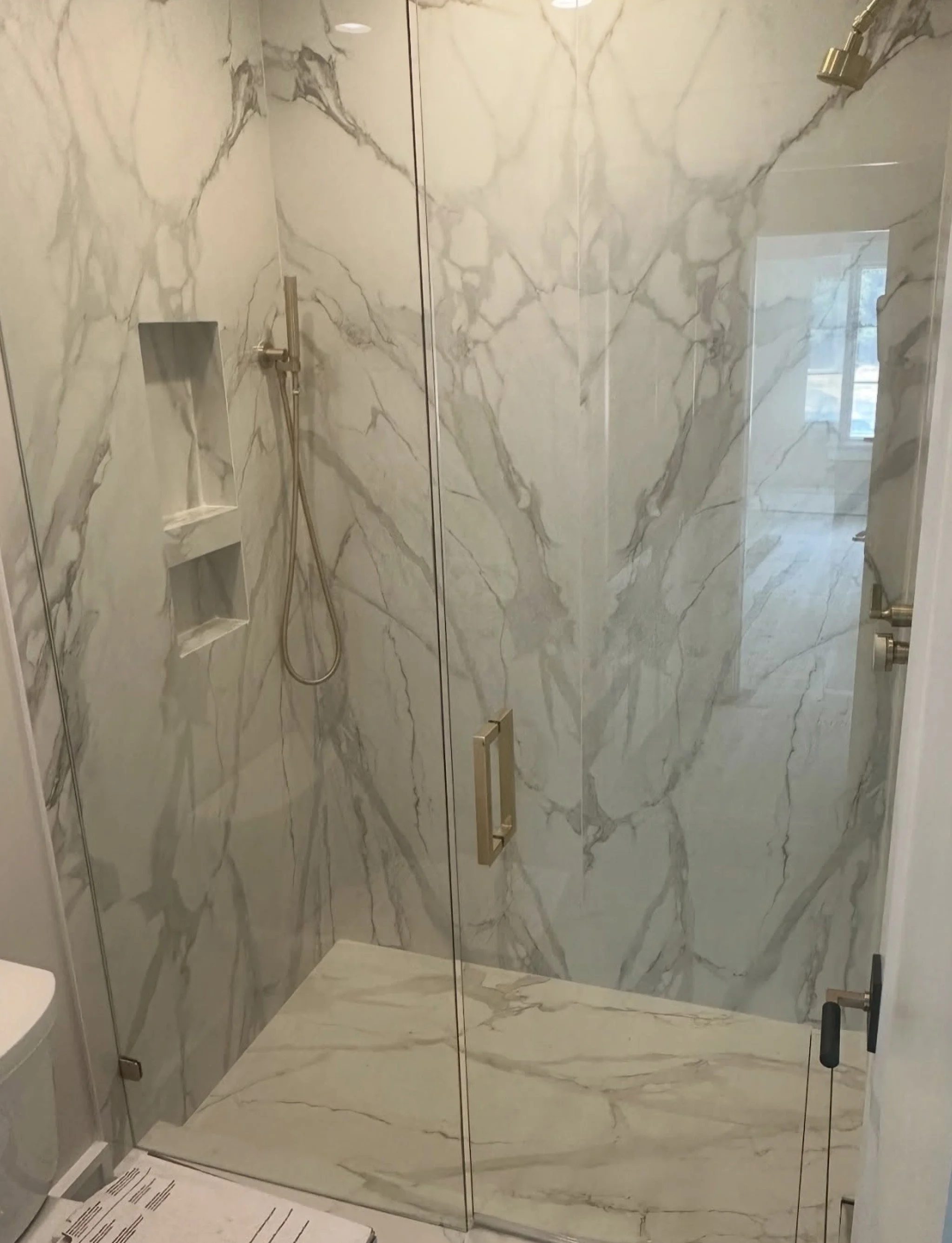 Bathroom shower with marble walls and floor, glass door, built-in shower shelves, and a handheld showerhead.
