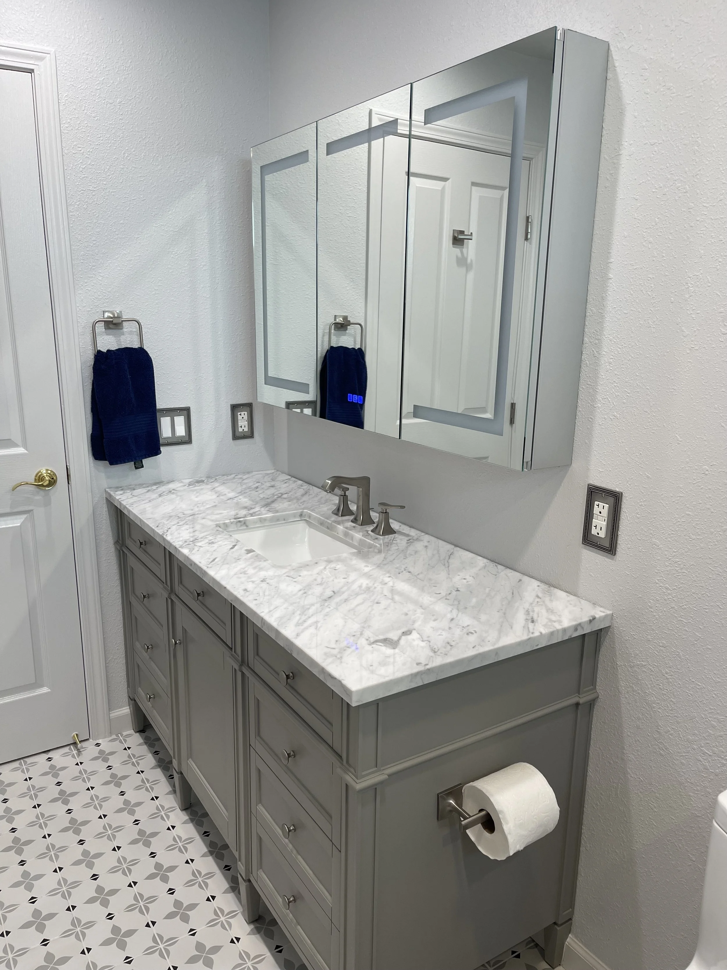 Bathroom vanity with gray cabinetry, marble countertop, and a large mirror with cabinet doors. There are two navy towels hanging on towel rings, a toilet paper roll on a holder, and patterned floor tiles.