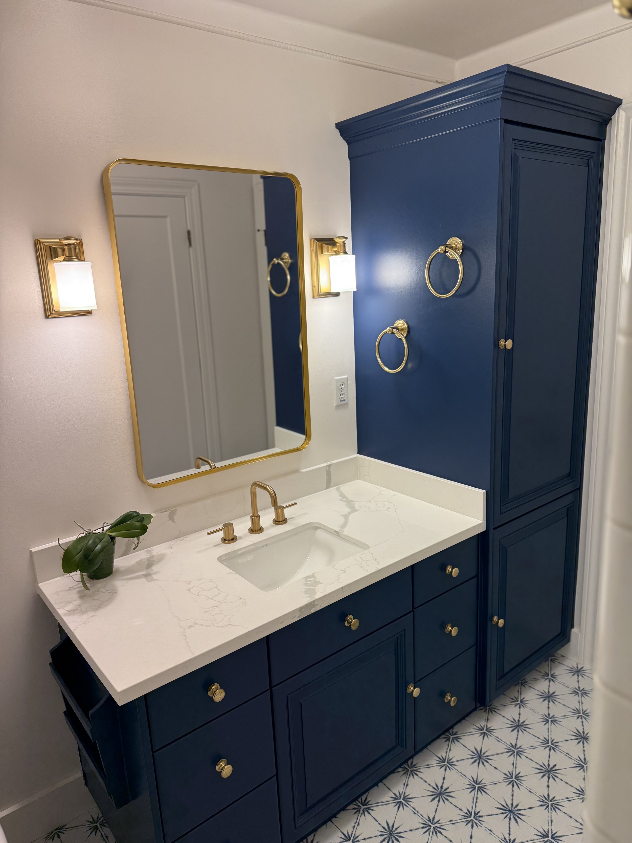 Blue bathroom vanity with gold accents, white marble countertop, and small plant on the left side. Mirror and two wall-mounted light fixtures with white shades above the vanity. Blue cabinet with gold handles adjacent to the vanity. Patterned tile fl