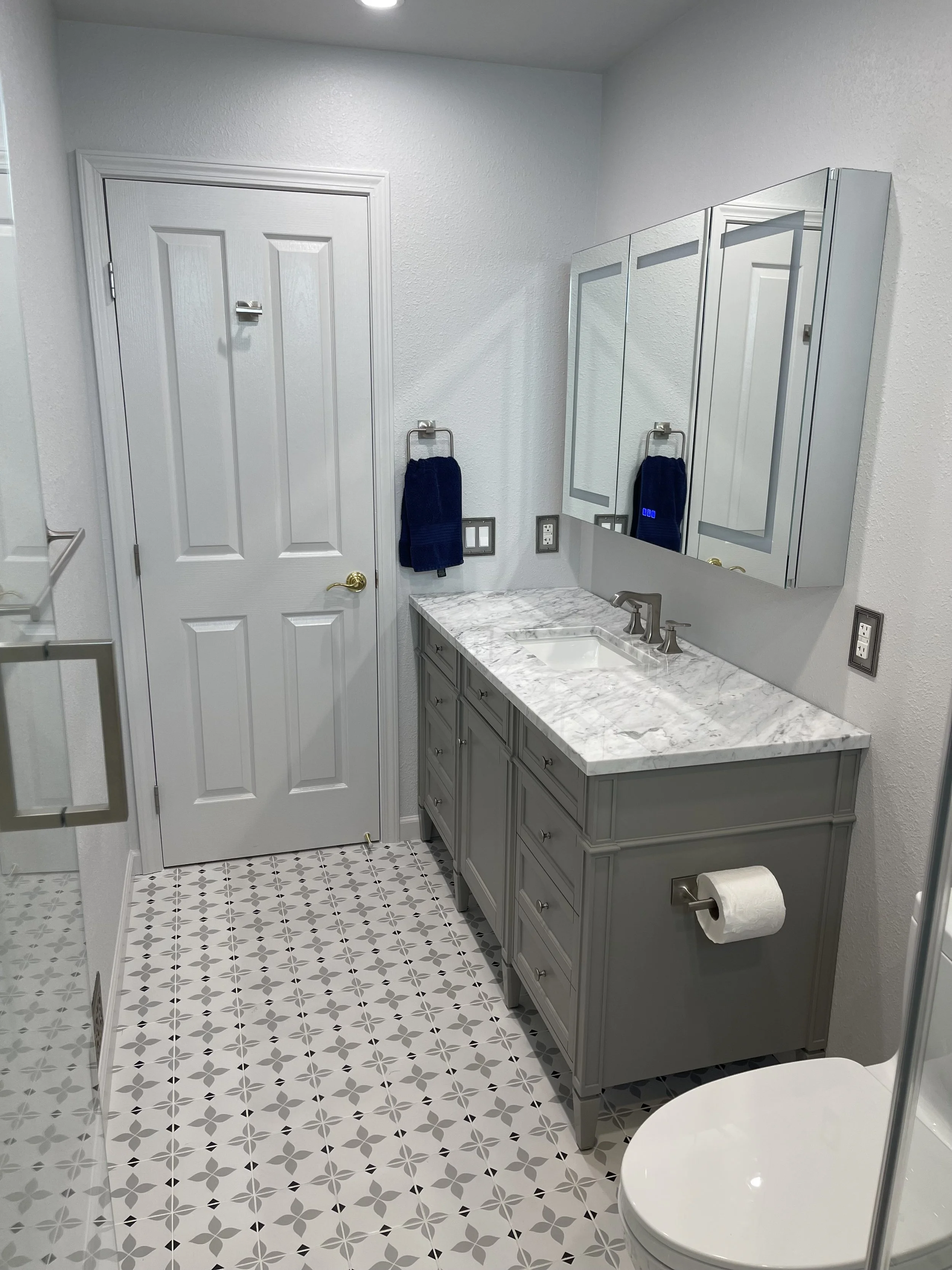 A small bathroom with a white door, grey vanity with a marble countertop, mirror cabinet, and a toilet with a roll of toilet paper. Two blue towels hang on hooks, and the floor has patterned tiles.
