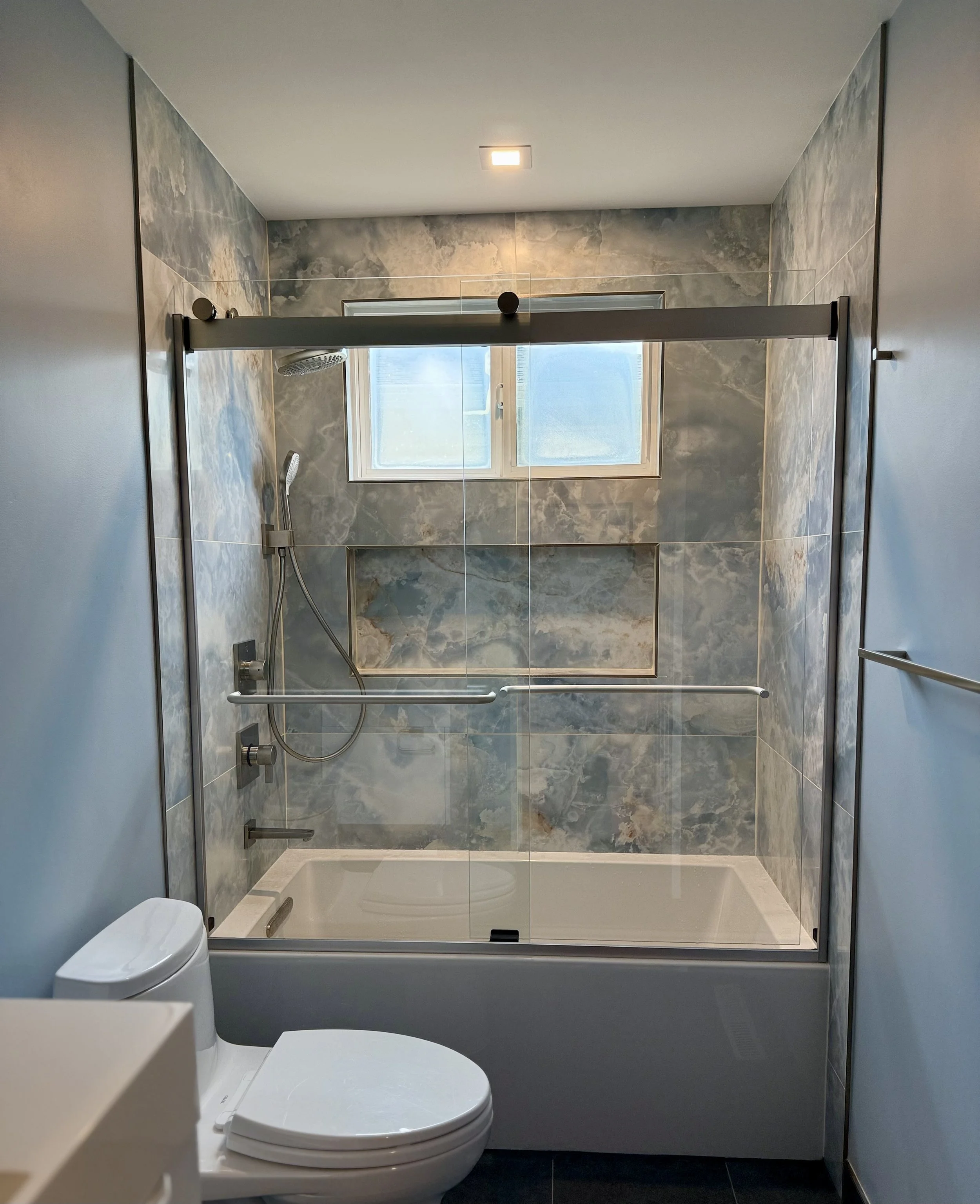 Bathroom with a walk-in shower with a sliding glass door, a window above the shower, marble tiles, and a toilet in the foreground.
