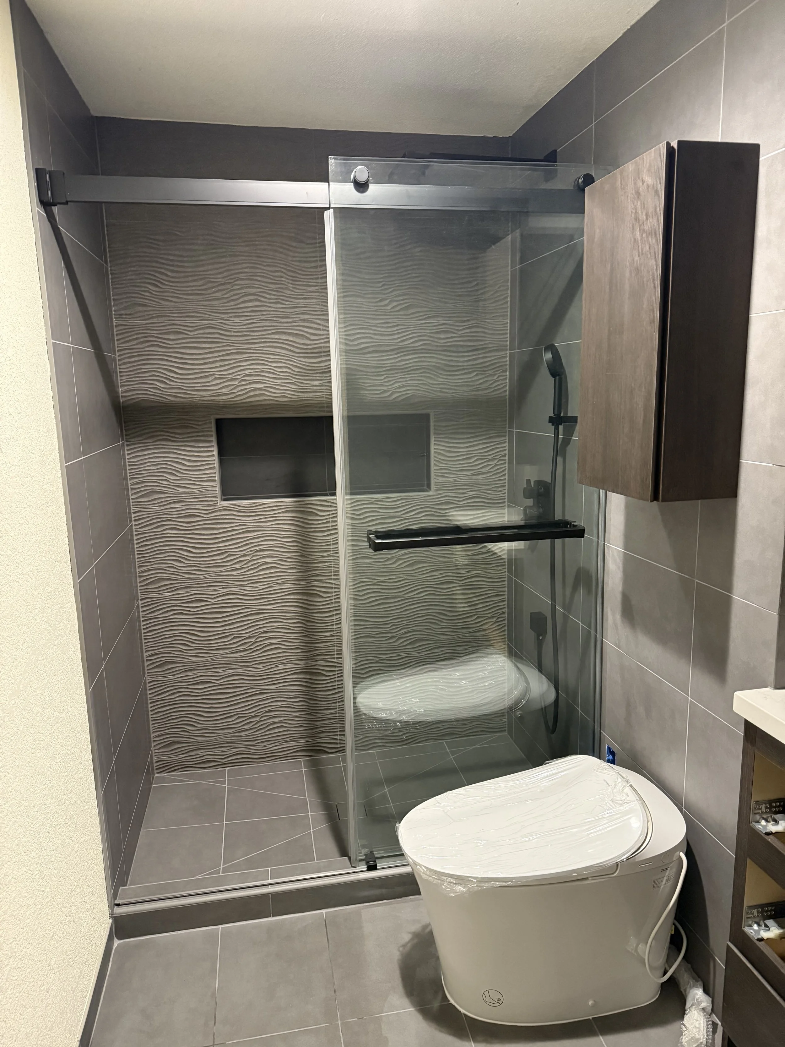 A modern bathroom with a tiled shower area, a glass door, a built-in shower niche, a handheld showerhead, a wooden wall-mounted cabinet, and a white smart toilet.