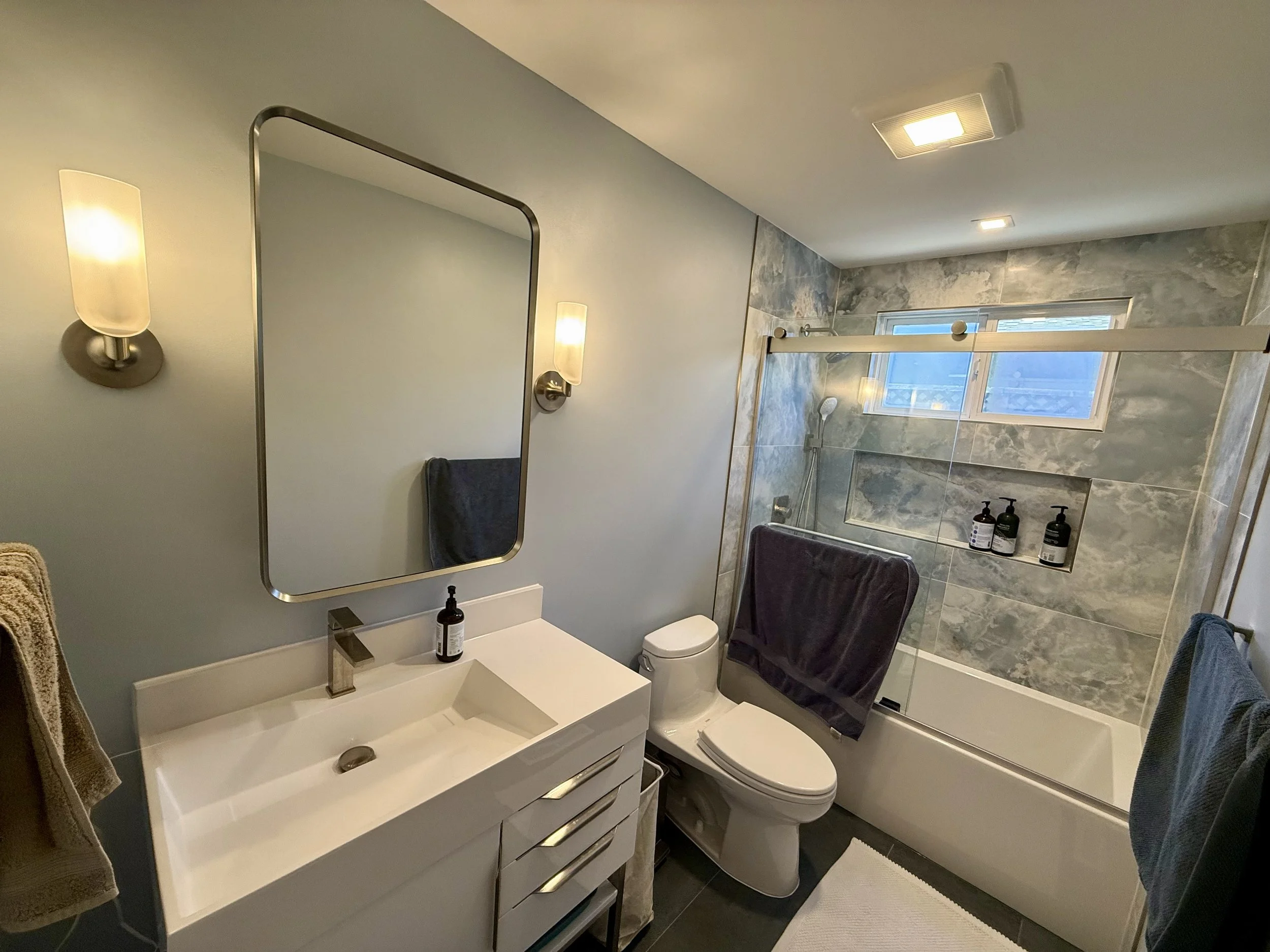 Modern bathroom with white vanity, rectangular mirror, wall-mounted light fixtures, toilet, and a bathtub with a sliding glass door. The shower area has gray marble tiles and a small window.