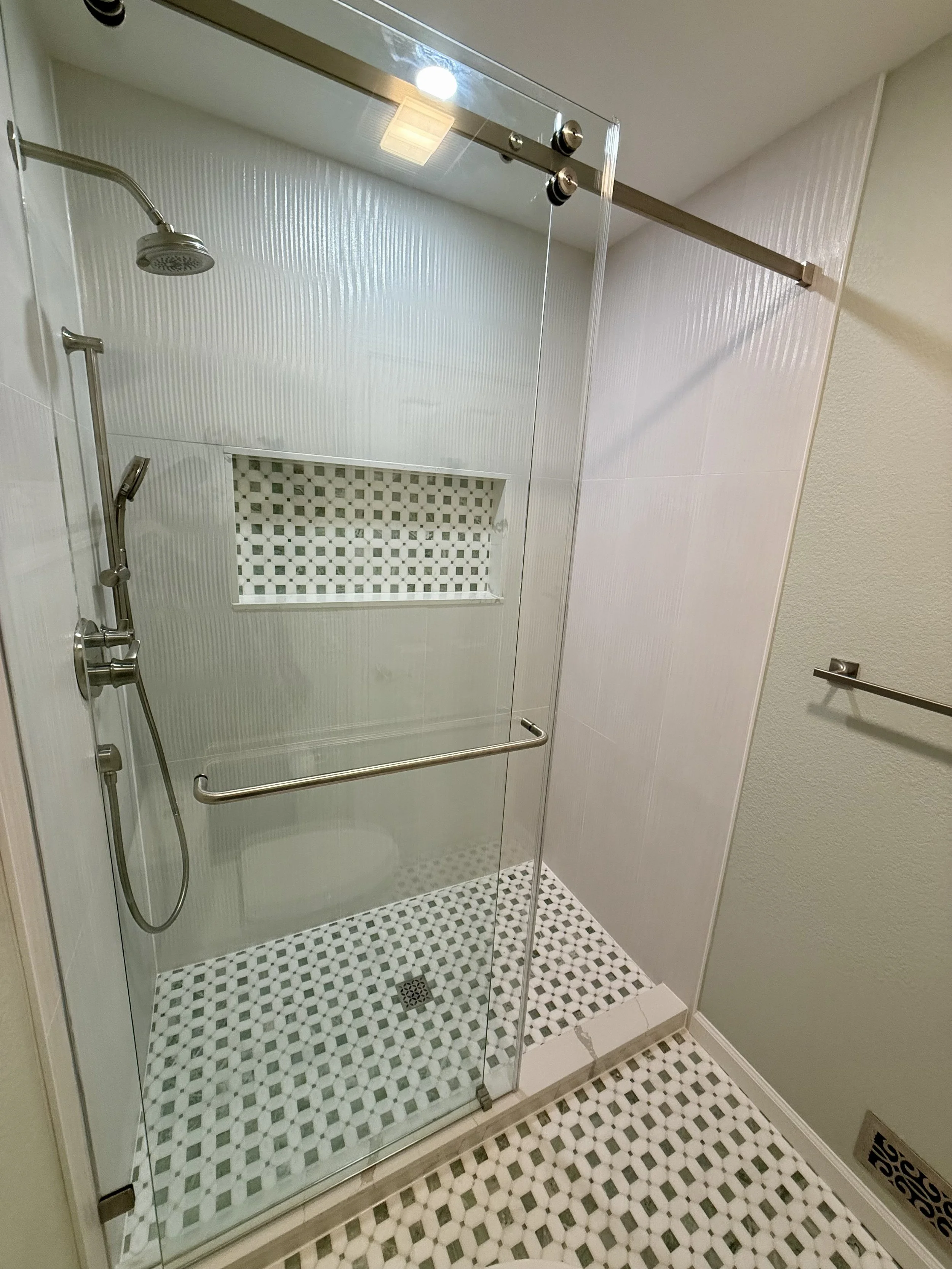 A glass shower enclosure with a metal sliding door, two shower heads, a built-in tiled niche, and a patterned tile floor and walls.