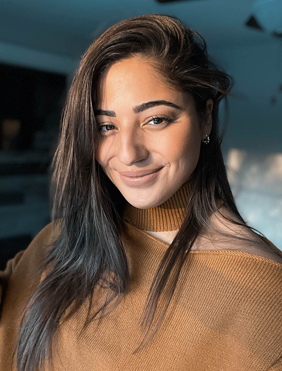 A young woman with long dark hair smiling, wearing a brown turtleneck sweater, in a warmly lit indoor setting.