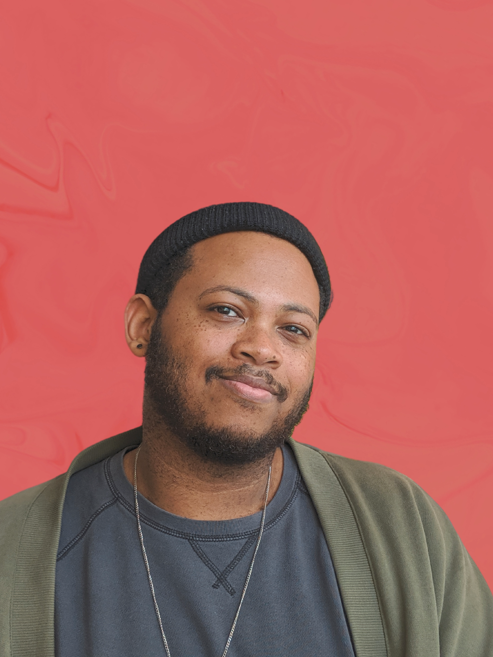 Portrait of a young man with a beard, wearing a black beanie, a gray T-shirt, and an olive green jacket, against a pink and orange marbled background.