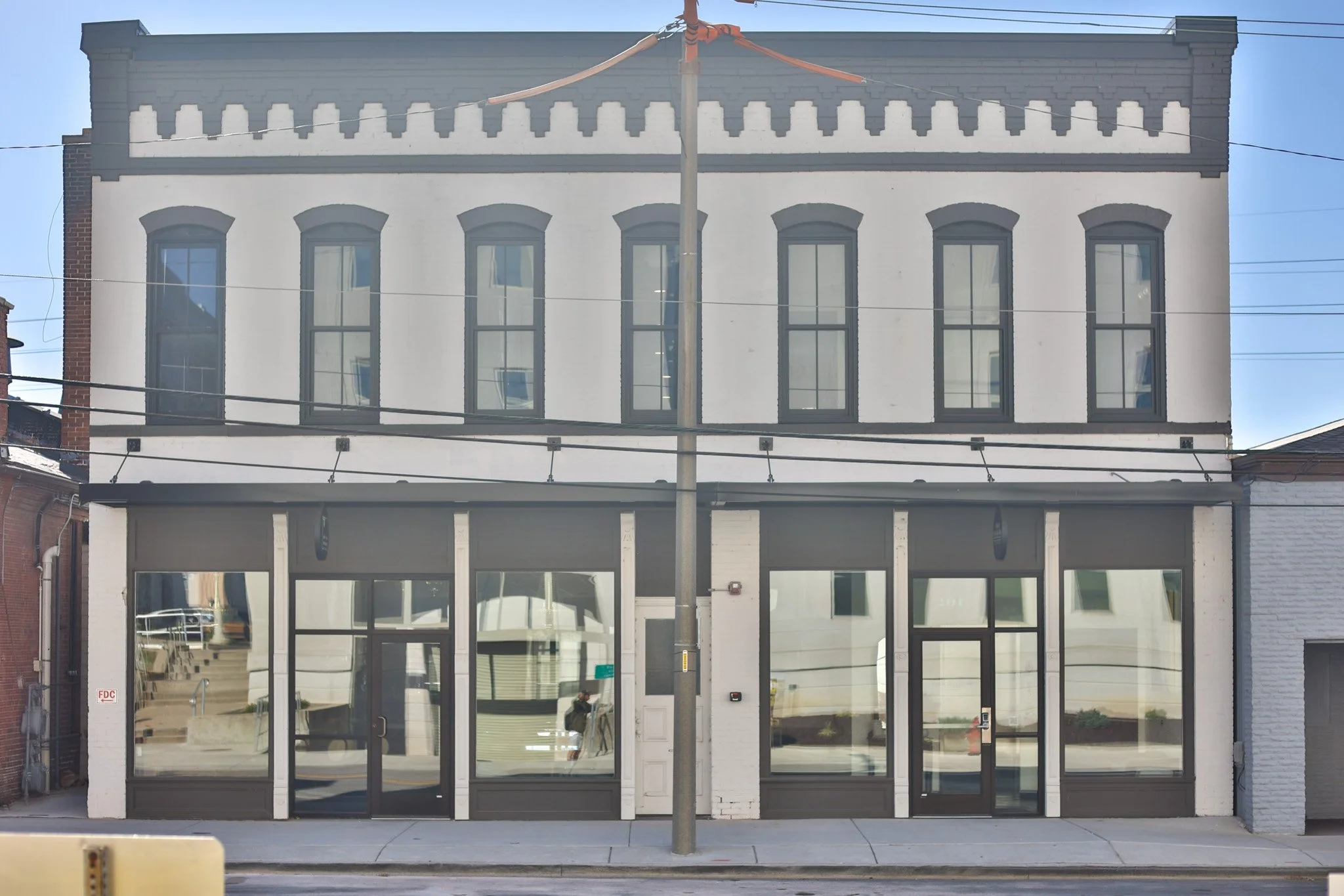 A three-story building with large glass windows and a white and gray exterior, with storefronts on the ground floor and windows on the upper floors, separated by a pole in the center.