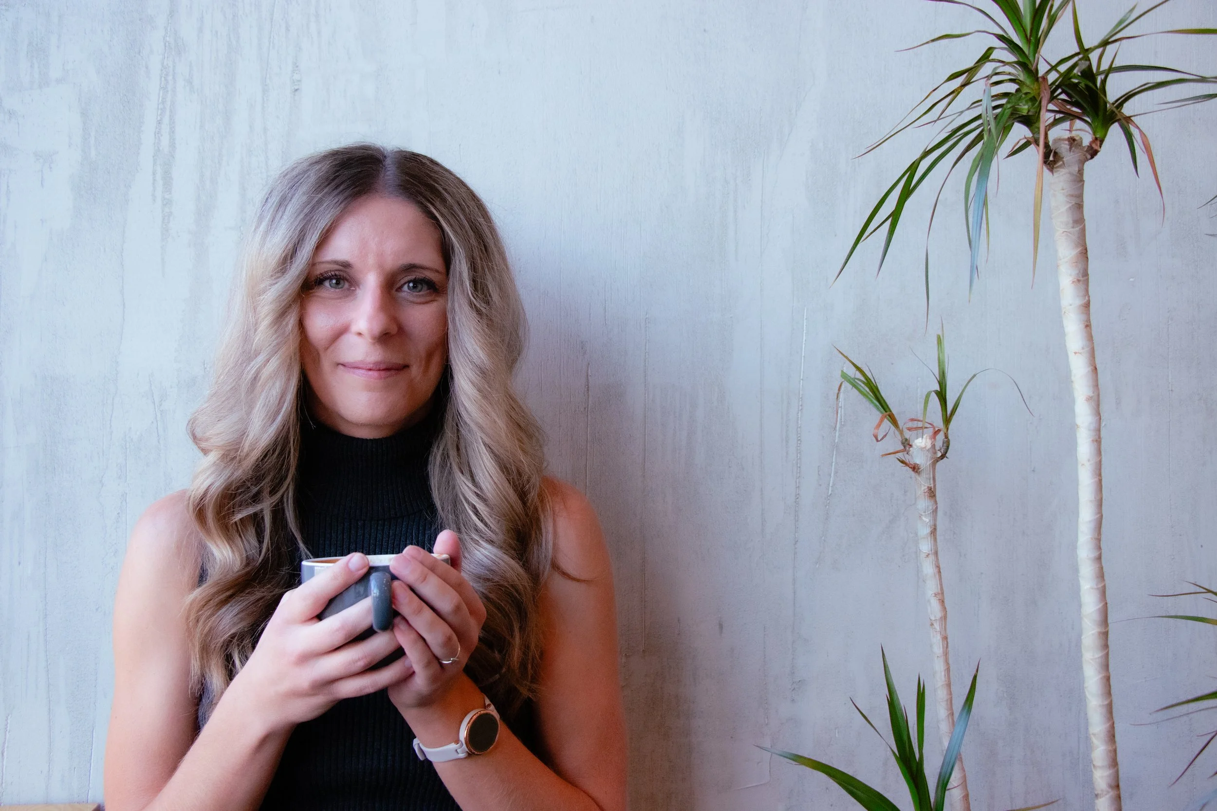 A woman with long wavy blonde hair wearing a black sleeveless turtleneck sweater and a white watch on her left wrist, holding a mug with both hands, standing next to a tall green plant with elongated leaves against a white textured wall.