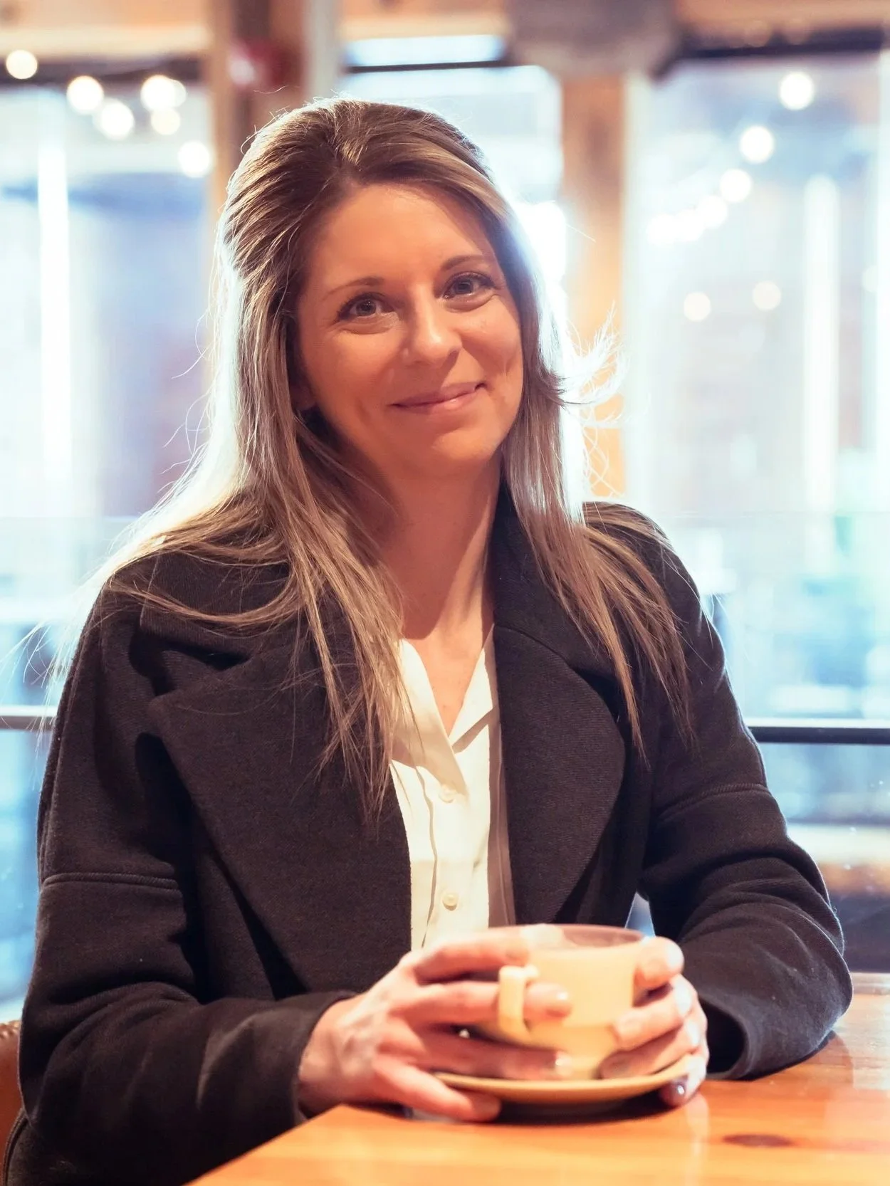A woman with long, light brown hair, smiling, sits in a café holding a beige coffee mug with both hands. She is wearing a black jacket and a white blouse. The background features blurred lights and windows with natural light coming through.