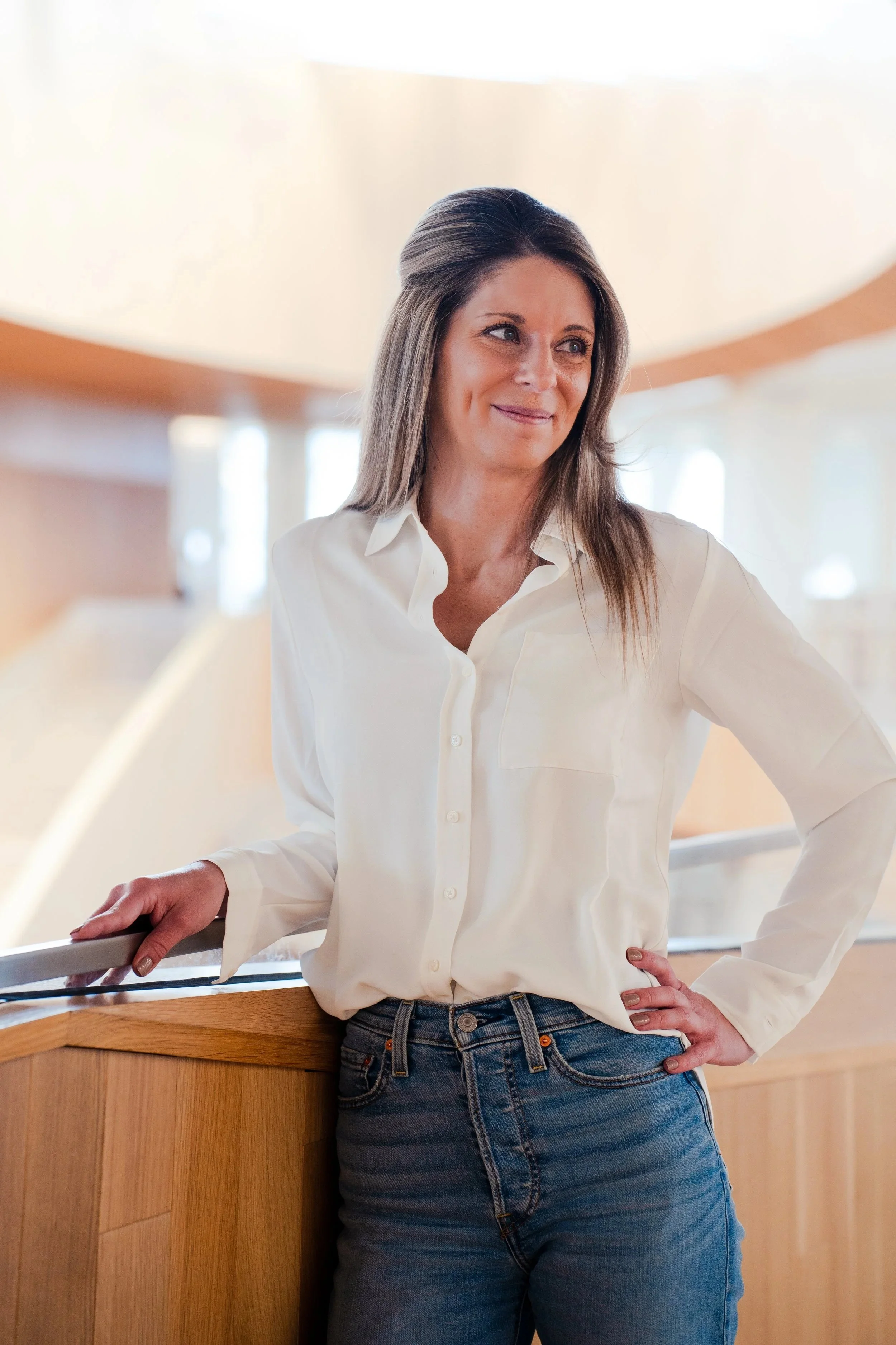 Woman in white shirt and jeans standing at a wooden counter, looking to her right, with her left hand resting on the counter and her right hand on her hip, in a bright indoor setting.