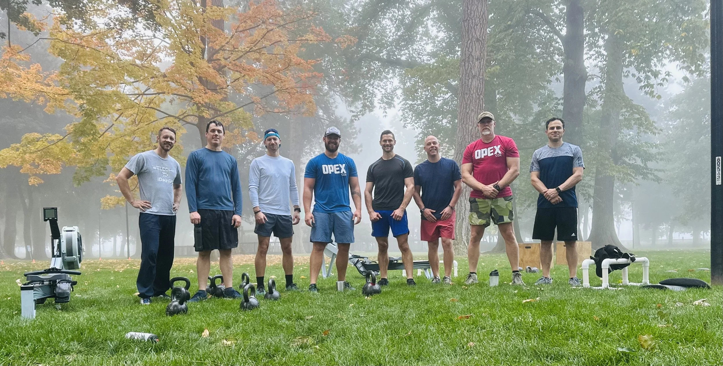 Group of nine men standing outdoors on grass, with various fitness equipment placed in front of them, including kettlebells, a rowing machine, and a bench, in a foggy park with trees in autumn.