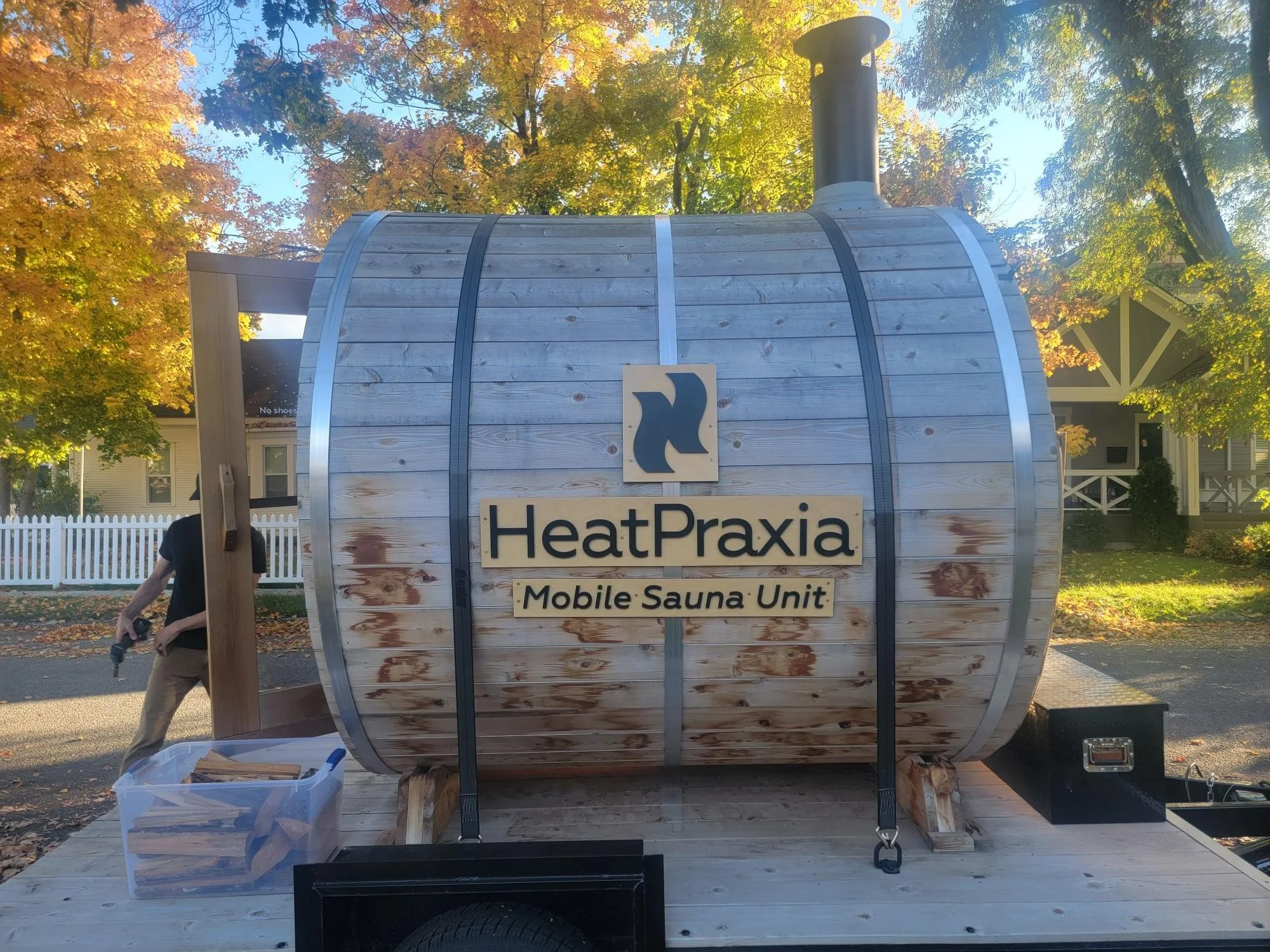 A mobile sauna unit named HeatPraxia, shaped like a large wooden barrel, is outdoors with trees and houses in the background. The sauna has a sign on it with a logo and the text 'HeatPraxia Mobile Sauna Unit'.