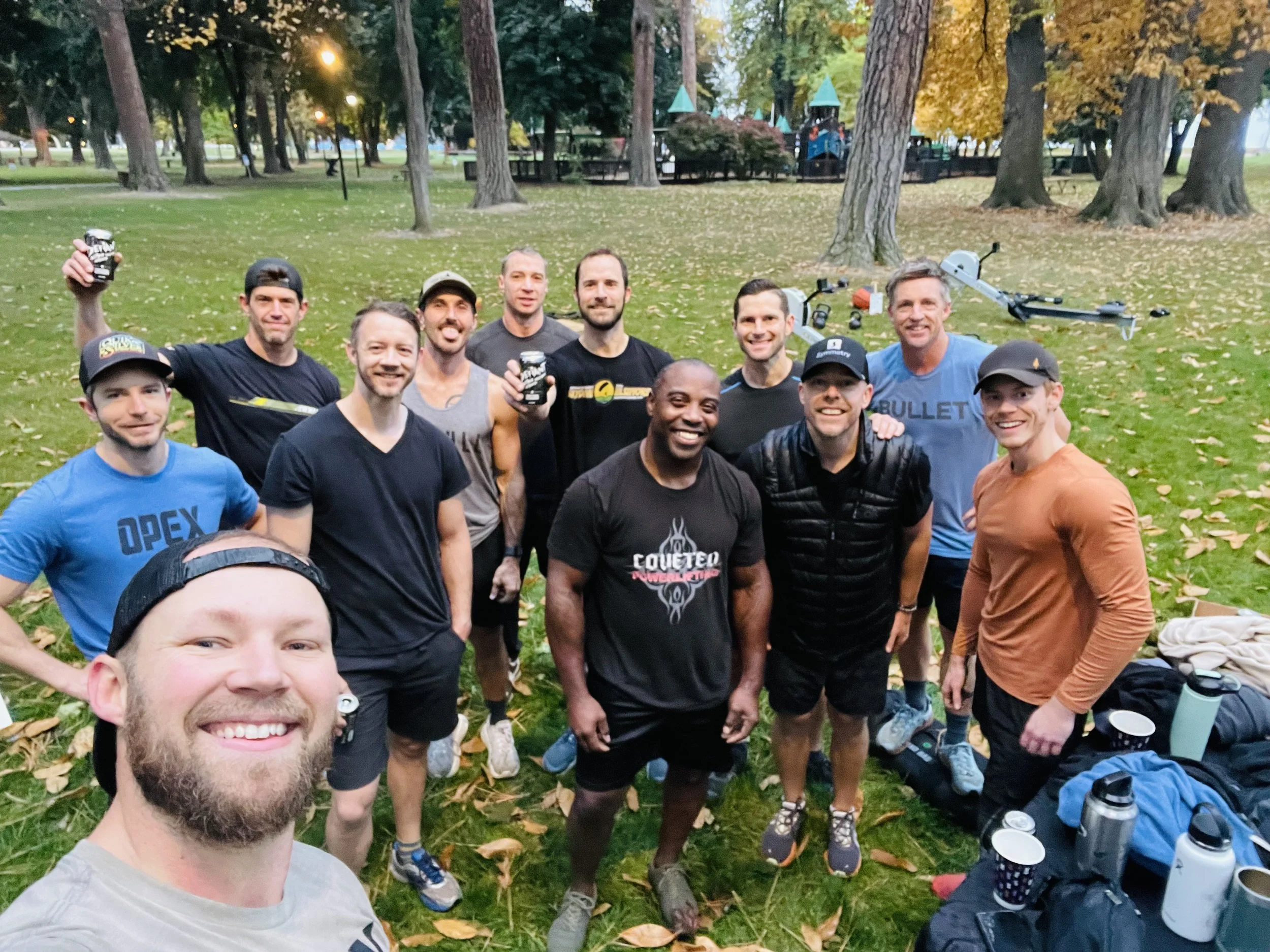 A group of people smiling and celebrating during a workout session in a park, with some holding drinks, and outdoor fitness equipment in the background.