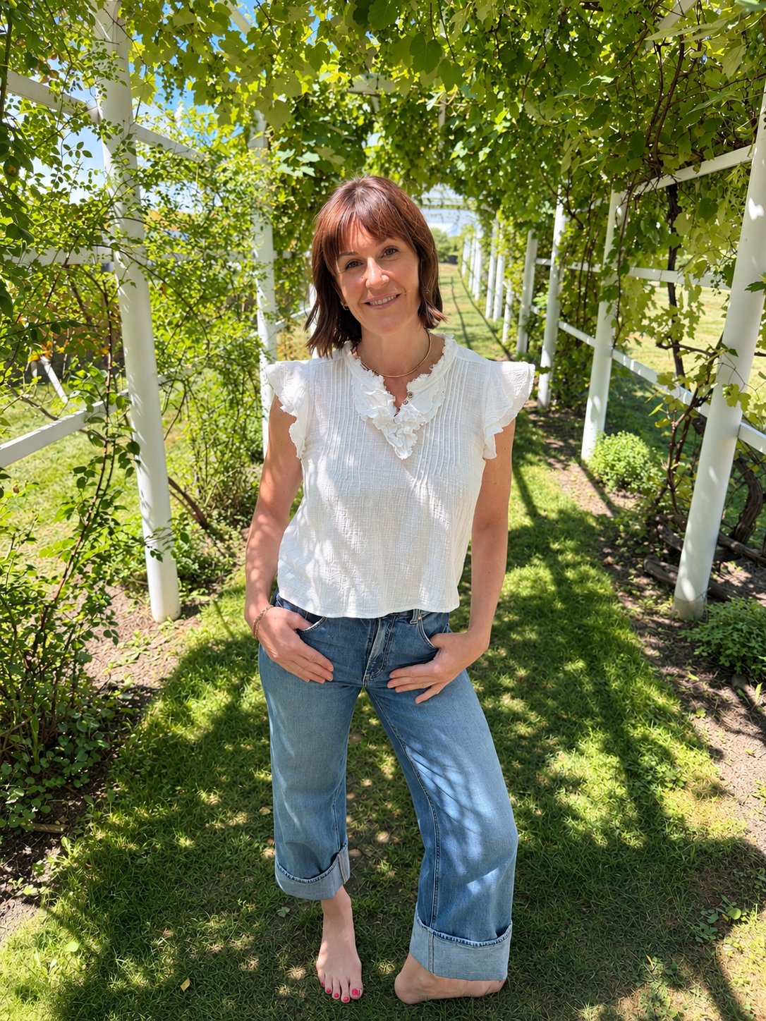 A woman with shoulder-length brown hair stands outdoors in a garden pathway surrounded by green vines and leaves, smiling at the camera, wearing a white blouse and blue jeans.