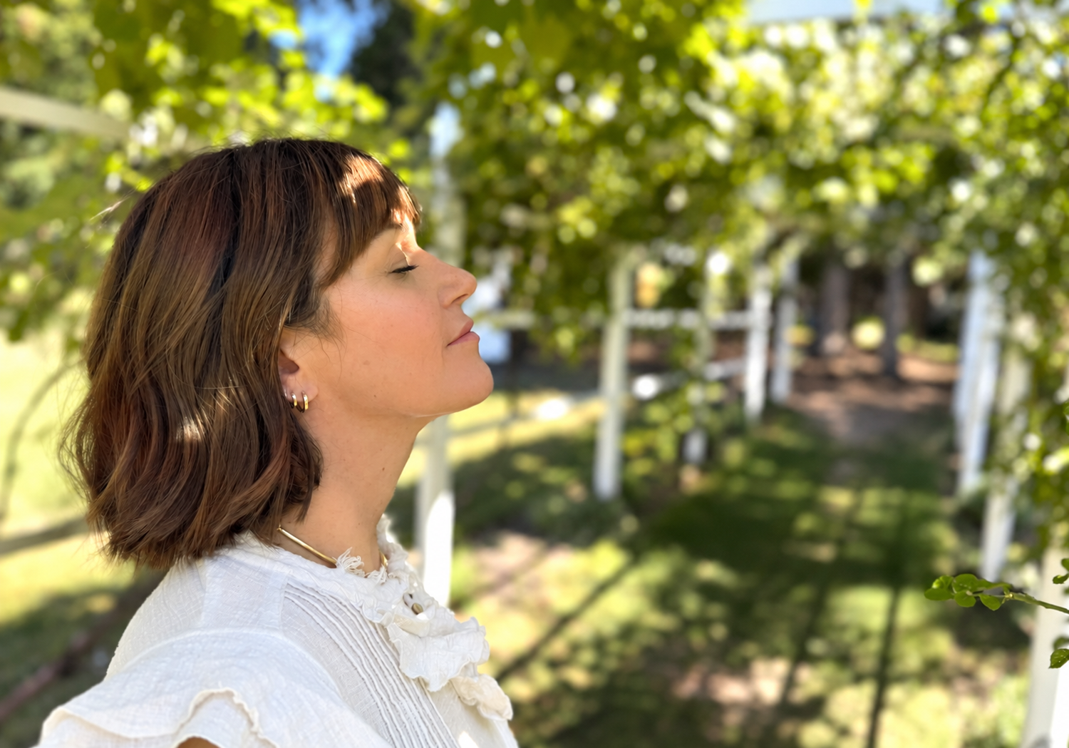 Side profile of a woman with short brown hair and earrings, standing outdoors with greenery and a white fence in the background.