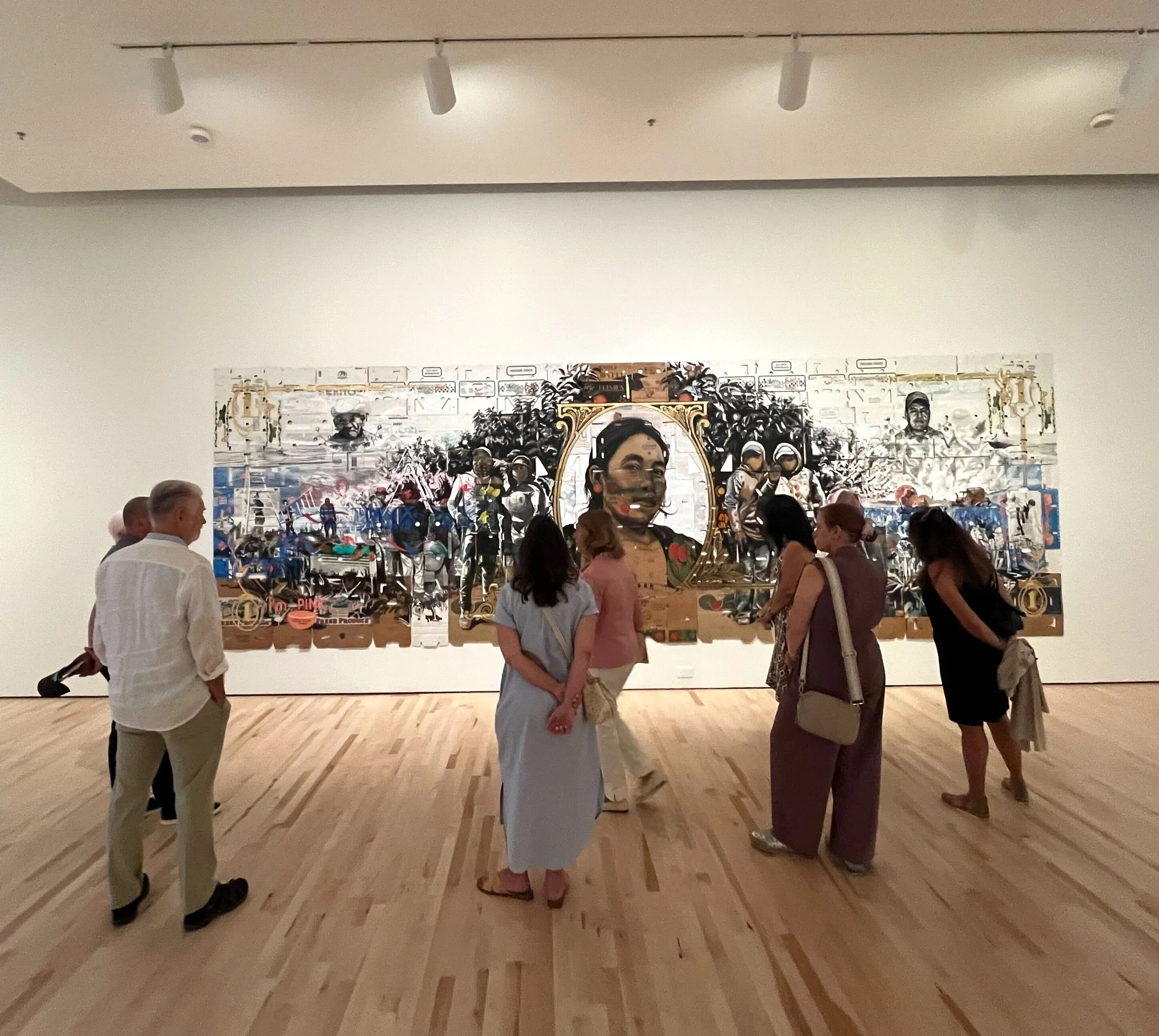 People viewing an artwork in a gallery, with a large mixed media painting featuring a woman's portrait surrounded by various scenes and figures.